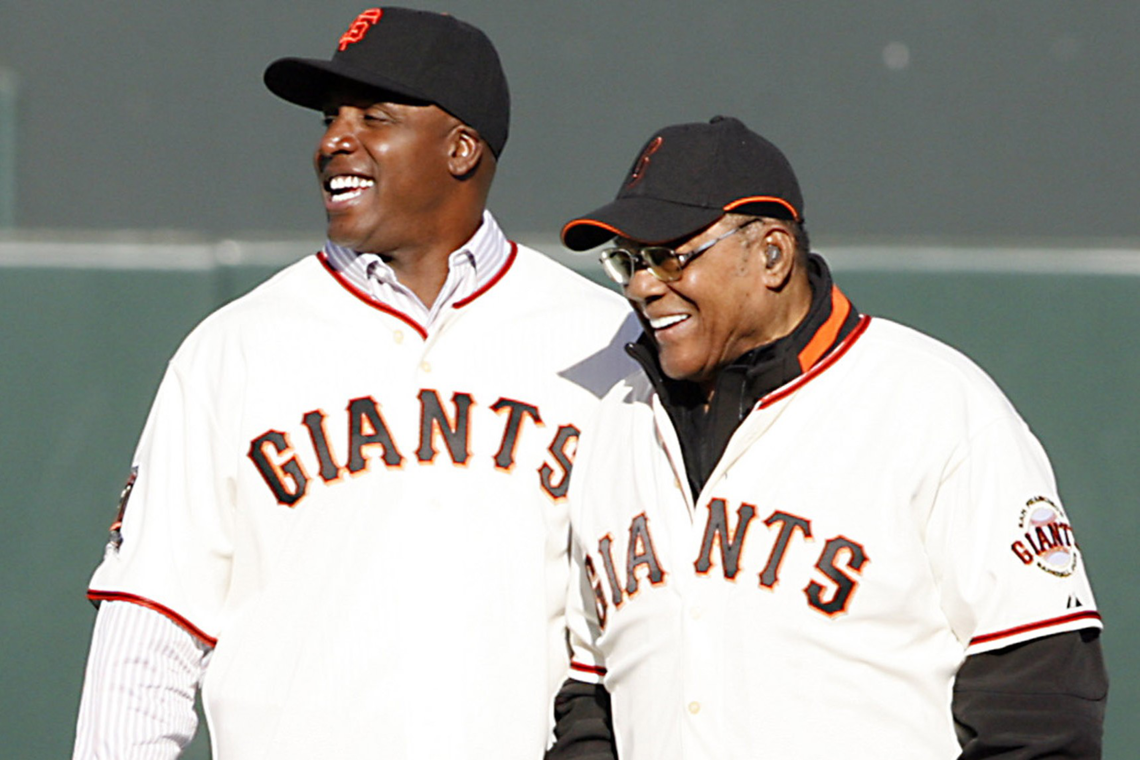 Two men wearing San Francisco Giants baseball jerseys and black caps are standing side by side, smiling and looking happy.