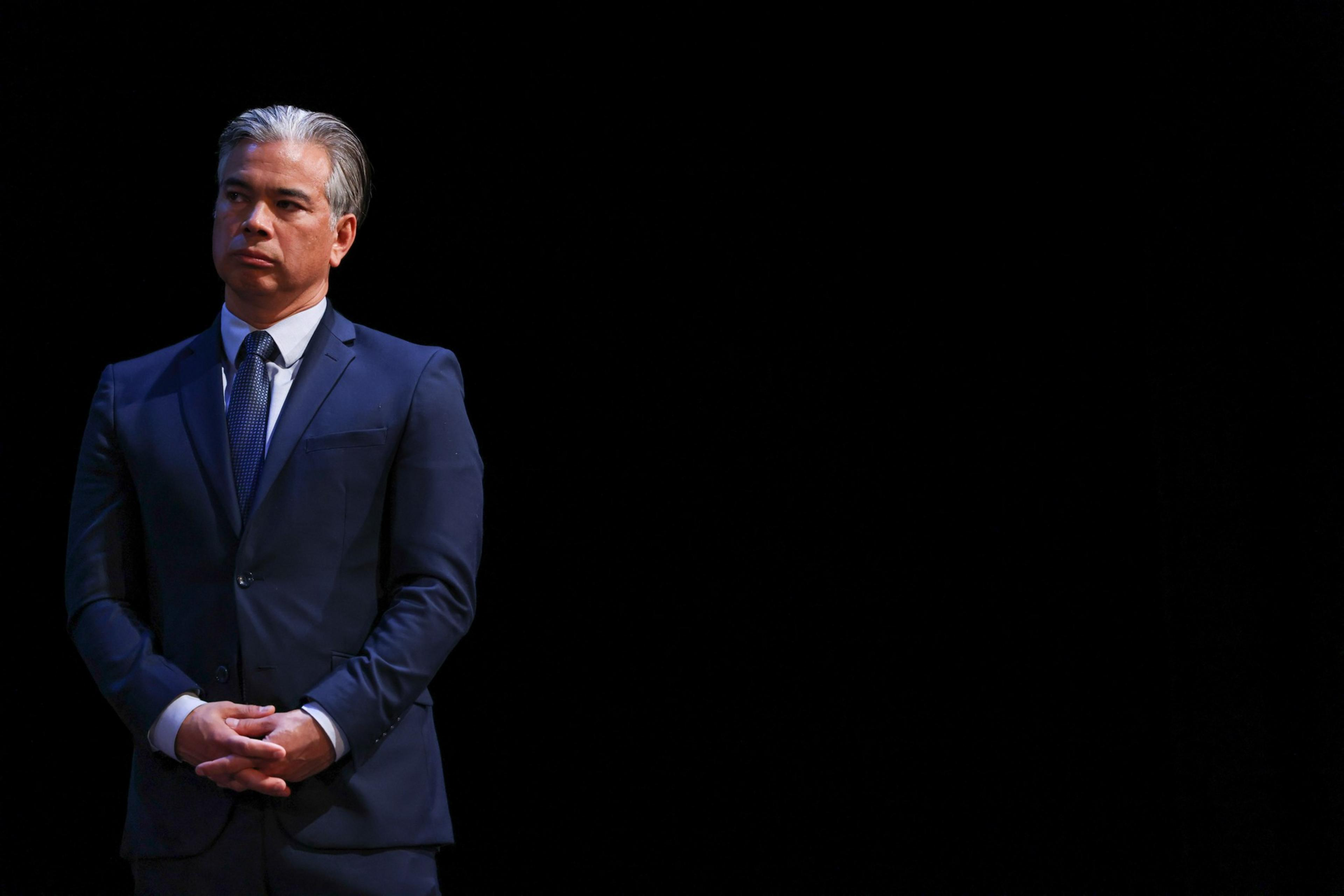 A man in a suit stands before a black background, looking pensive.