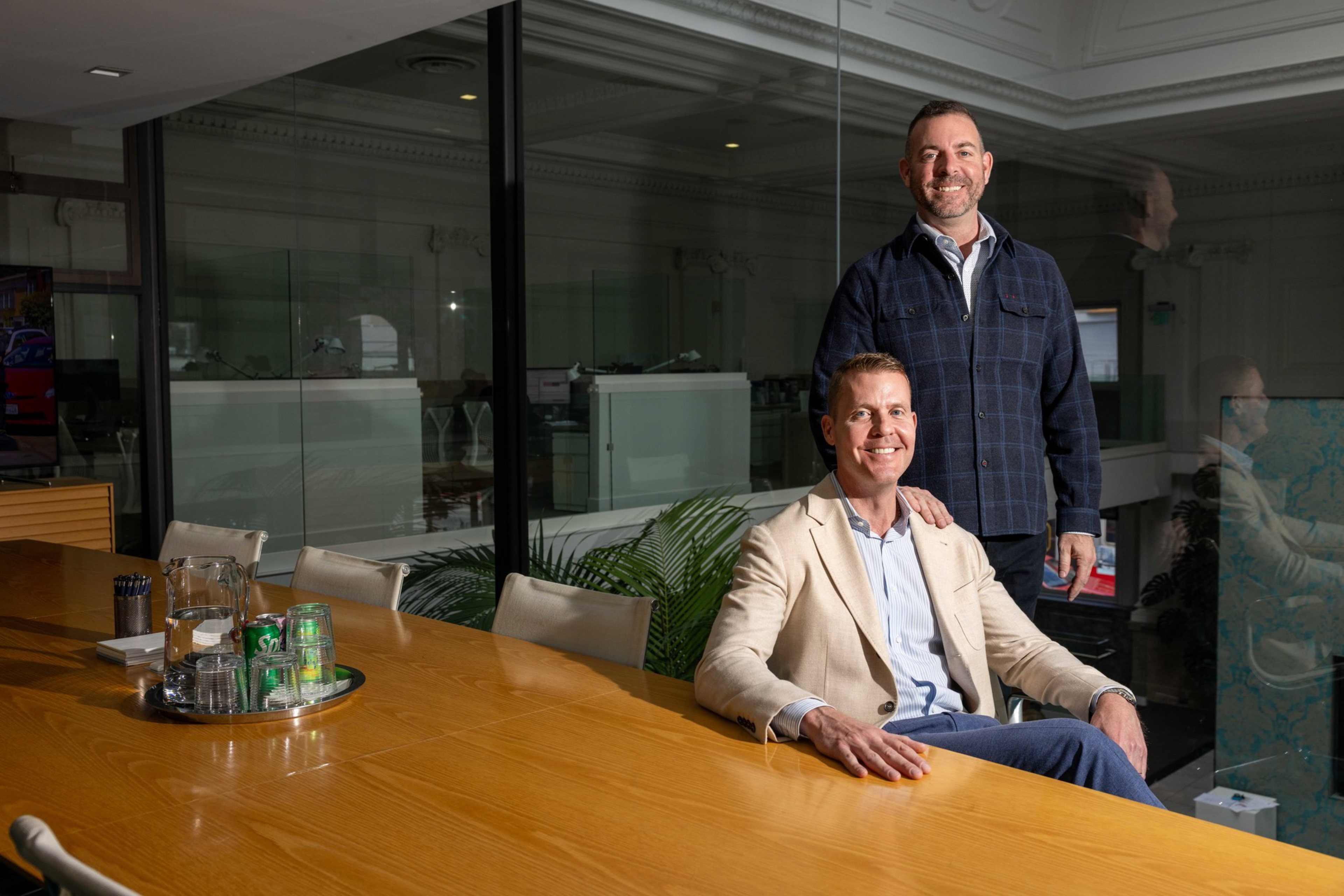 A man in a beige blazer sits at a conference table while another man in a dark jacket stands smiling behind him, with glass walls in the background.
