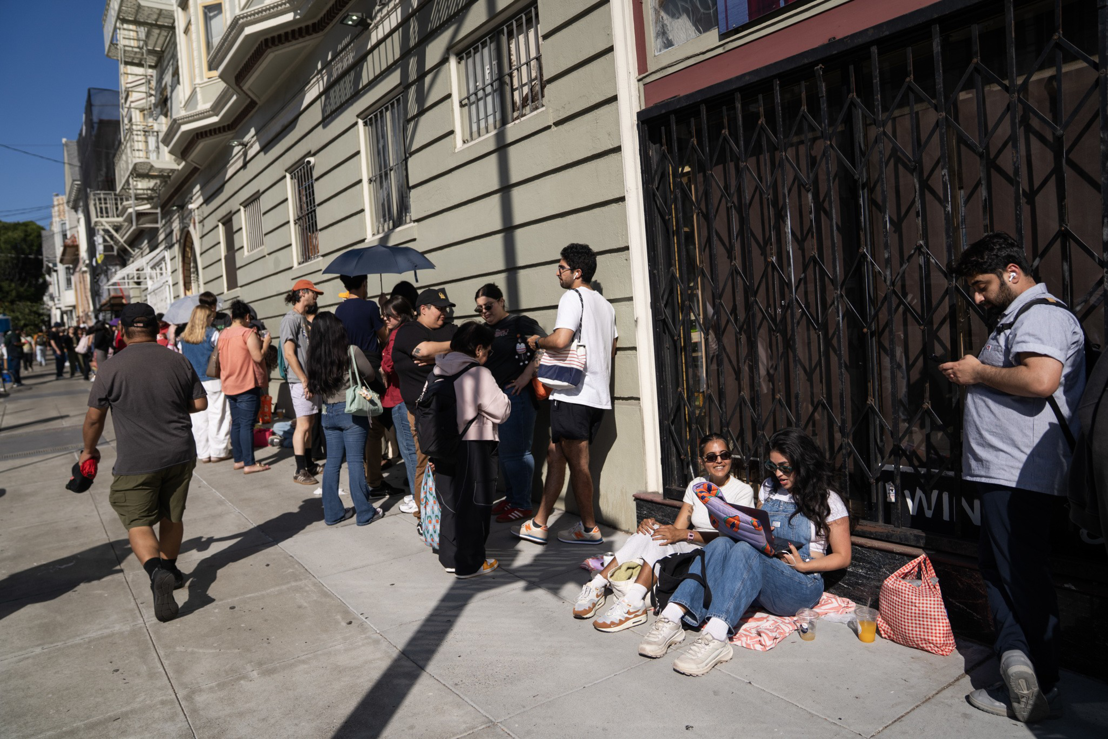 People stand in a long line along a city sidewalk, with two women sitting on the ground reading a flyer and a man looking at his phone nearby.