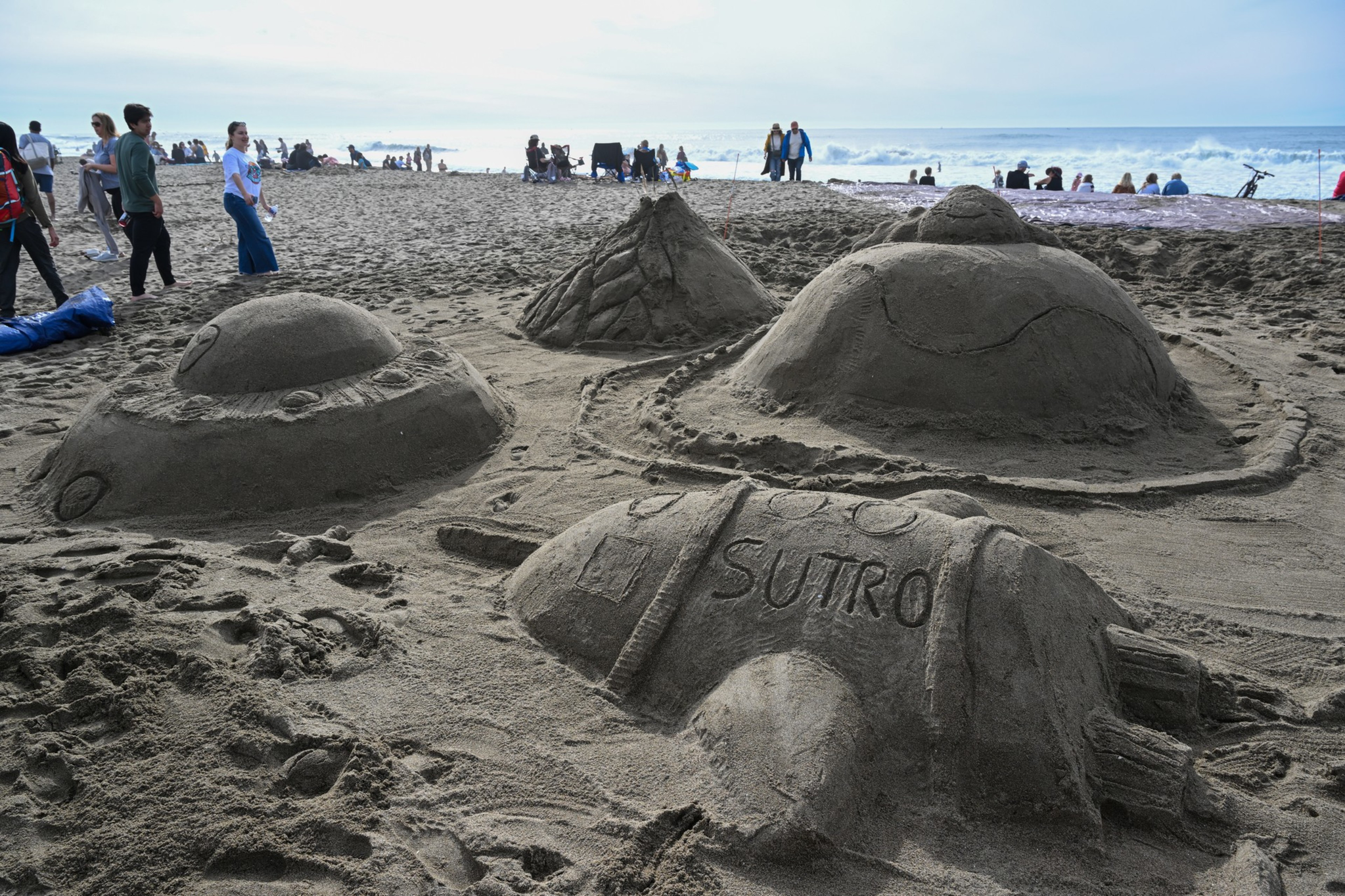 Four detailed sand sculptures resembling UFOs and a rocket labeled "SUTRO" sit on a sandy beach with people and ocean waves in the background.