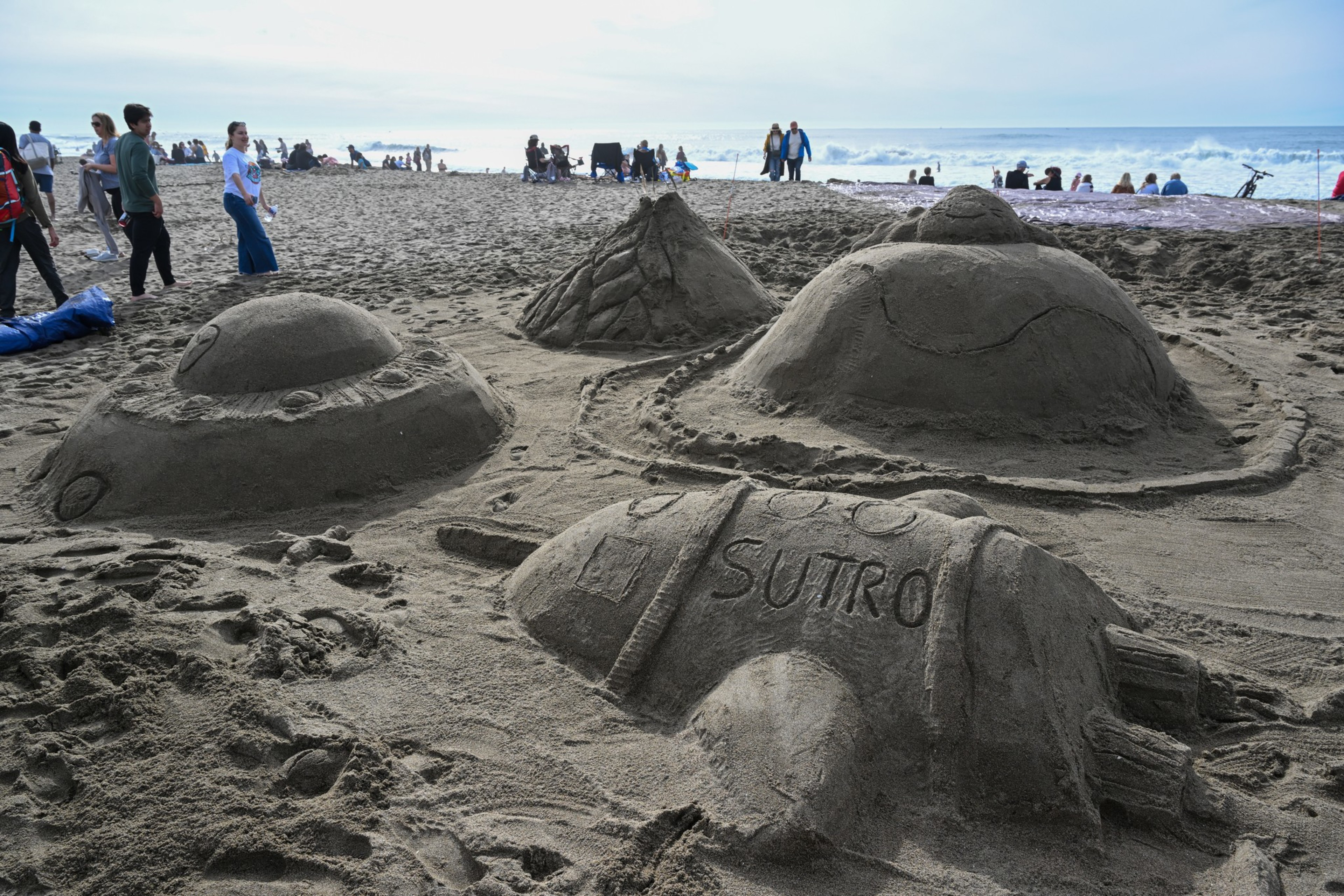 Four detailed sand sculptures resembling UFOs and a rocket labeled "SUTRO" sit on a sandy beach with people and ocean waves in the background.