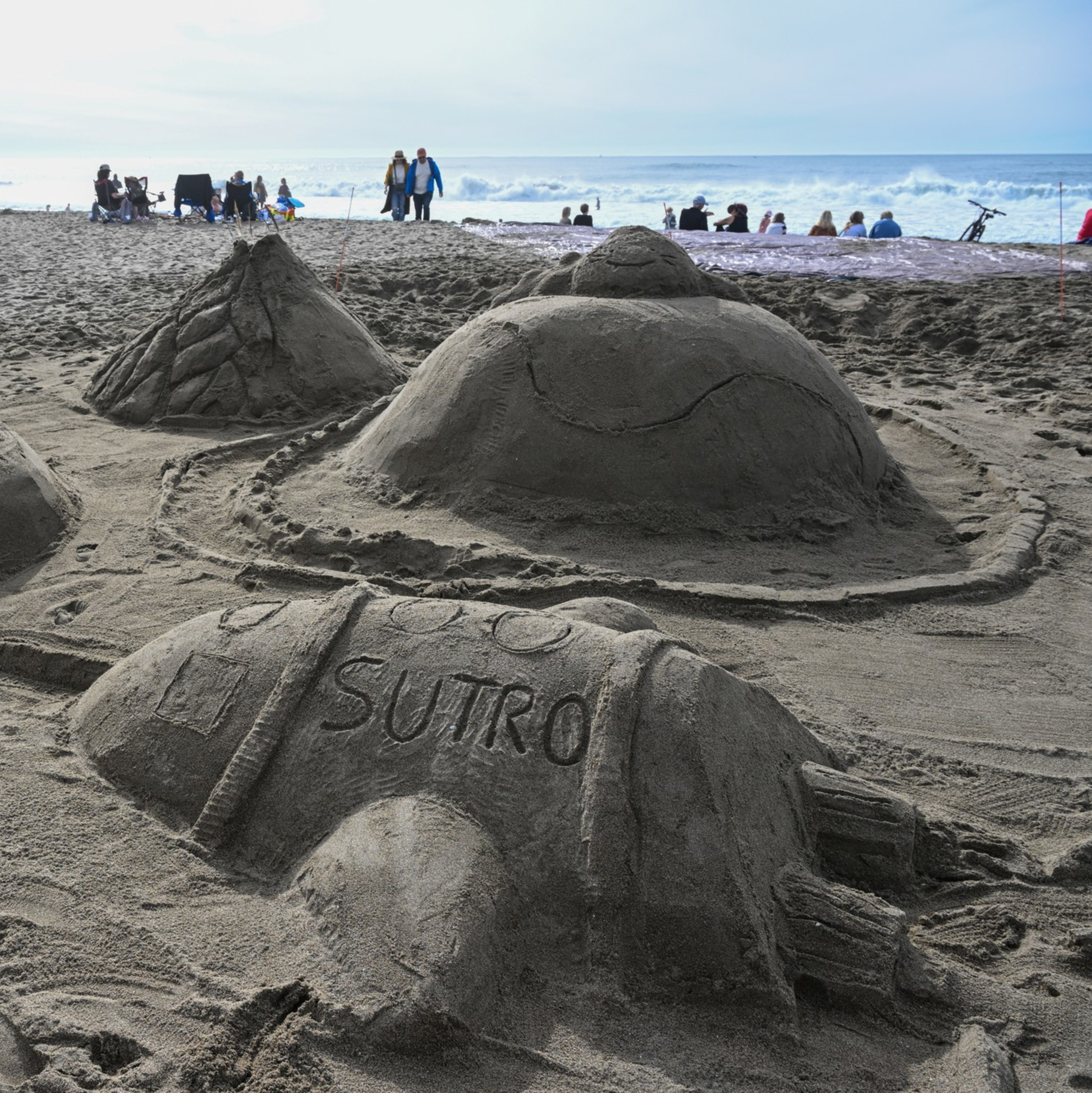 Four detailed sand sculptures resembling UFOs and a rocket labeled "SUTRO" sit on a sandy beach with people and ocean waves in the background.