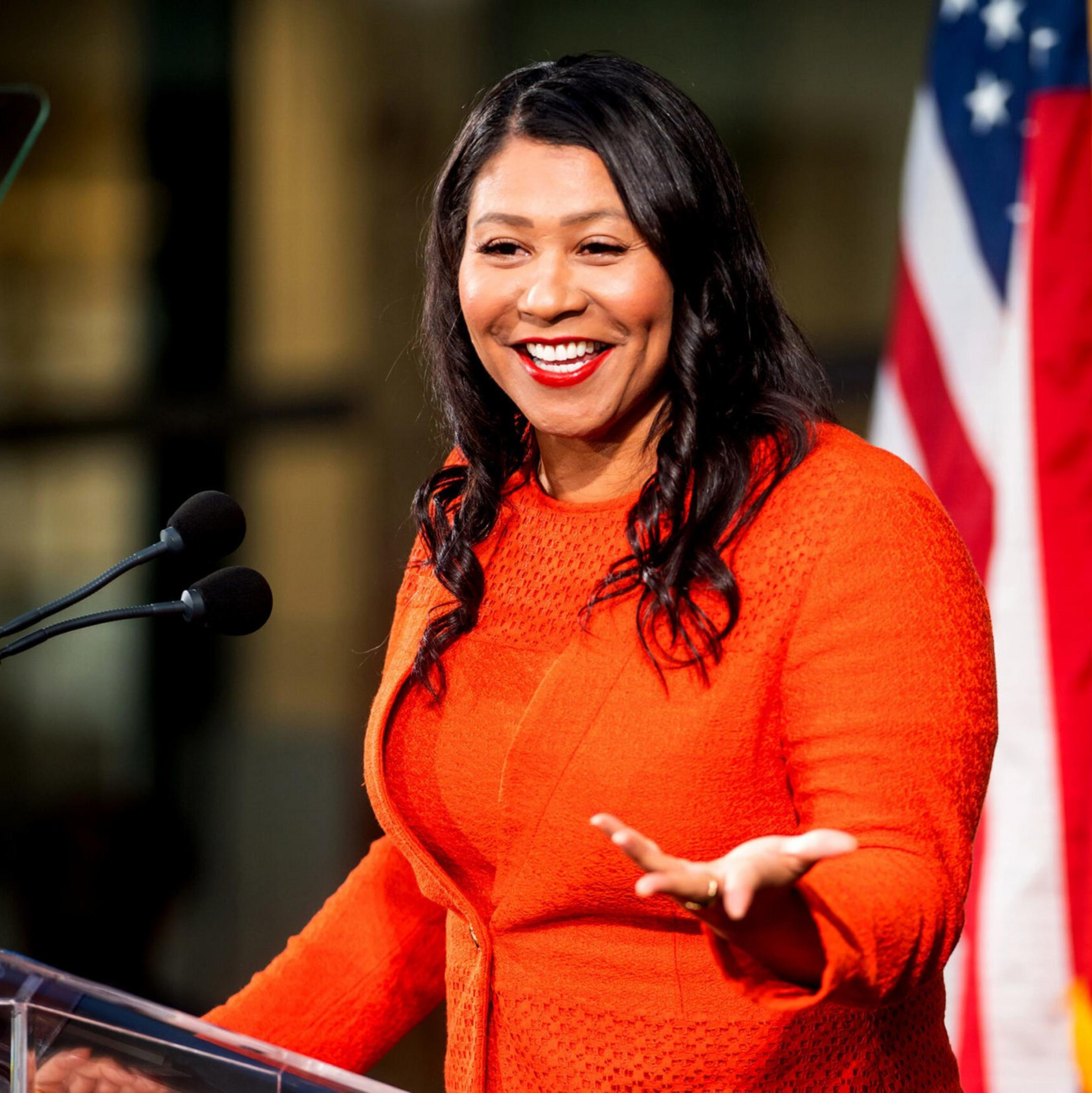 A woman in an orange dress smiles and gestures while speaking at a podium, with American and California flags in the background.