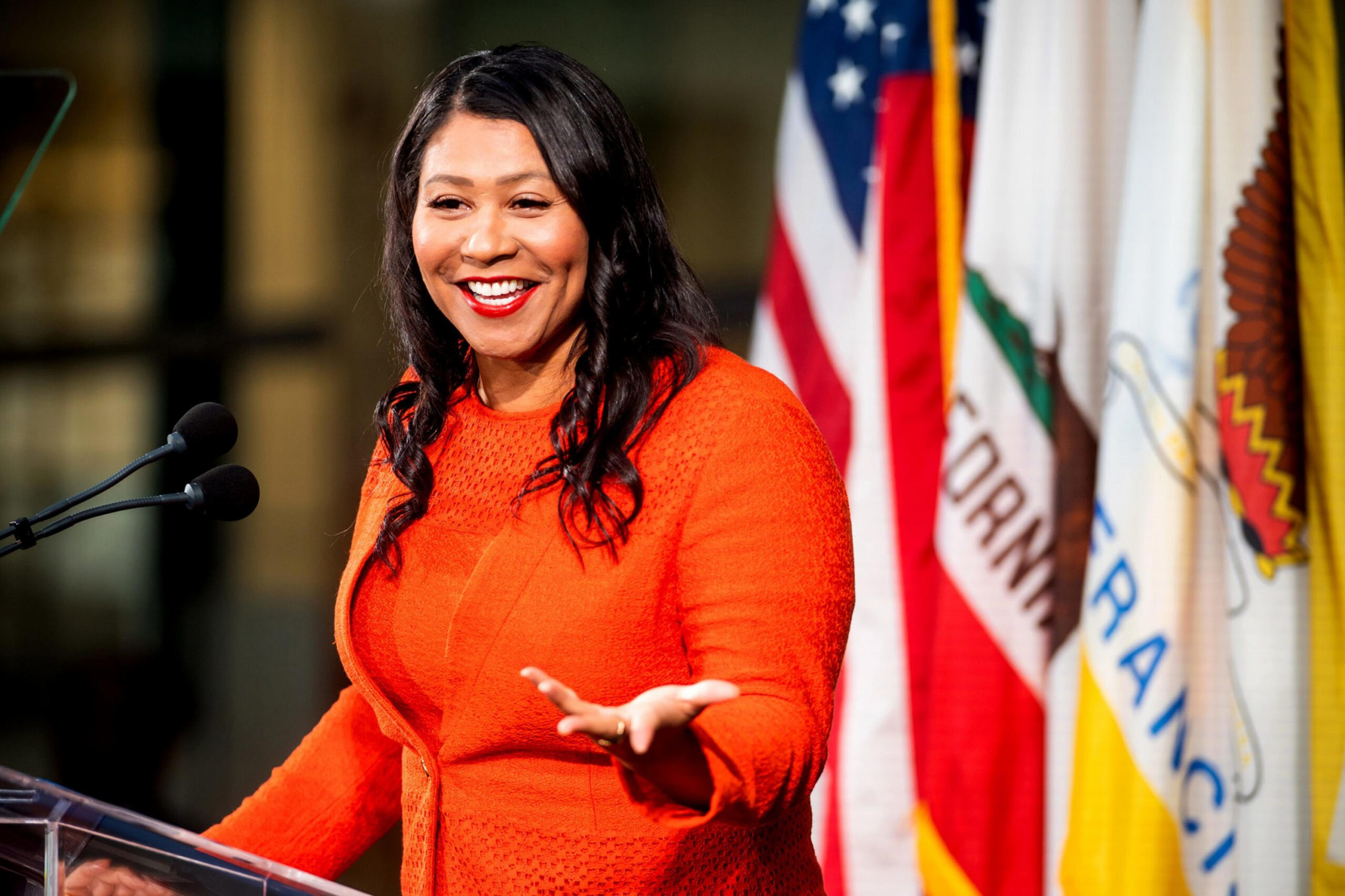 A woman in an orange dress smiles and gestures while speaking at a podium, with American and California flags in the background.