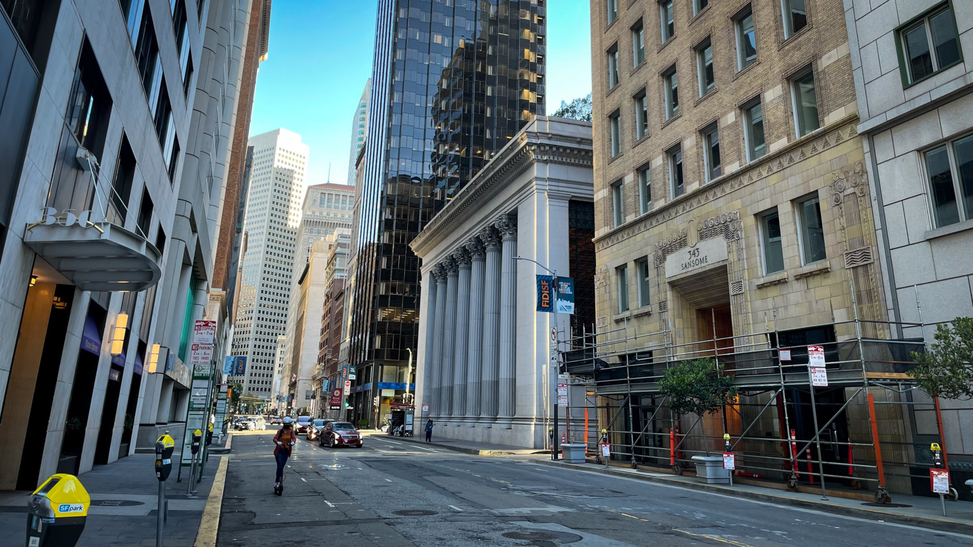 A wide city street lined with tall office buildings, including one with large columns, scaffolding on one side, and a few cars and a pedestrian.