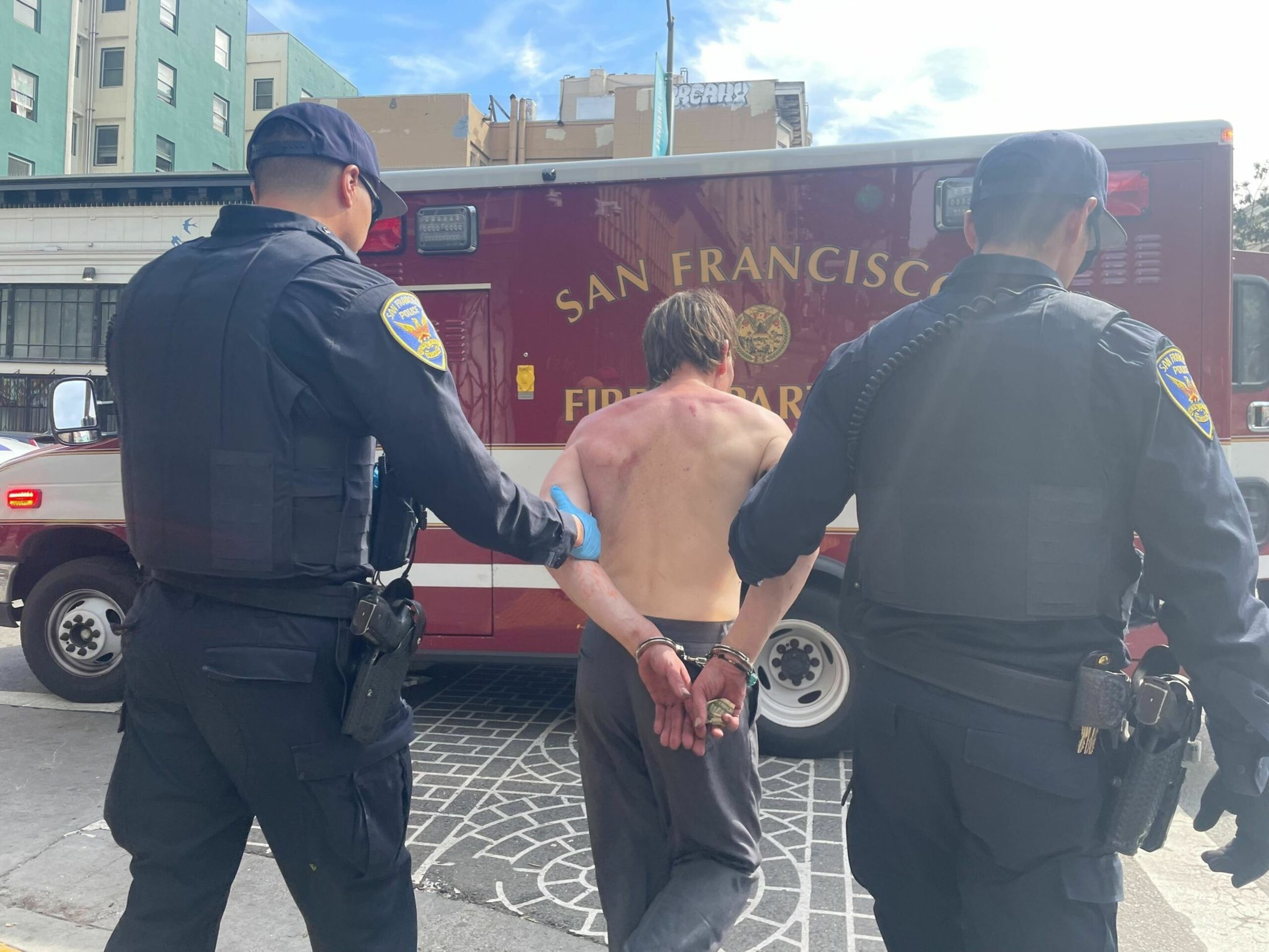 Two police officers escort a shirtless man in handcuffs toward a San Francisco Fire Department vehicle.