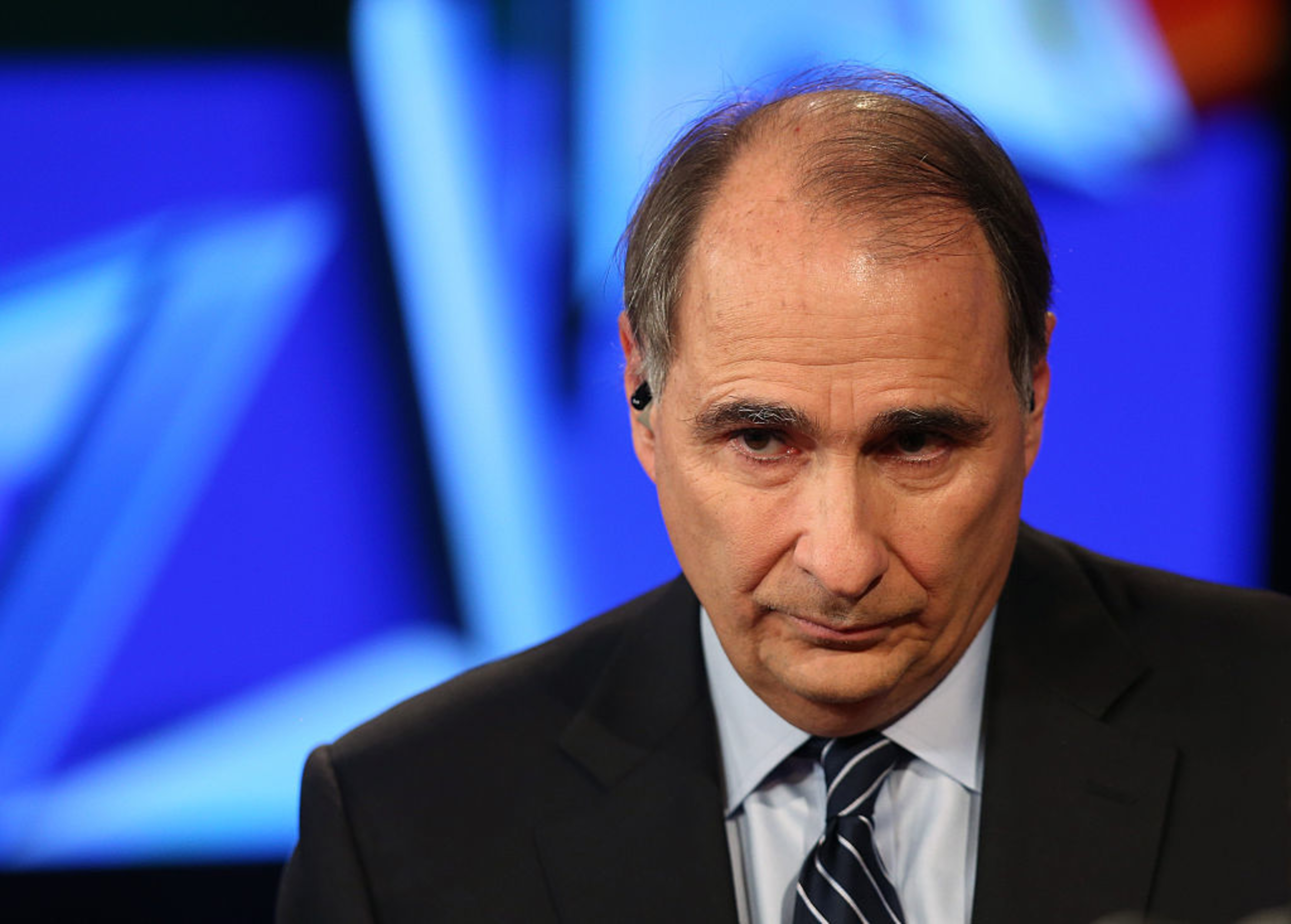 A middle-aged man in a dark suit and striped tie looks serious against a blurred blue background.