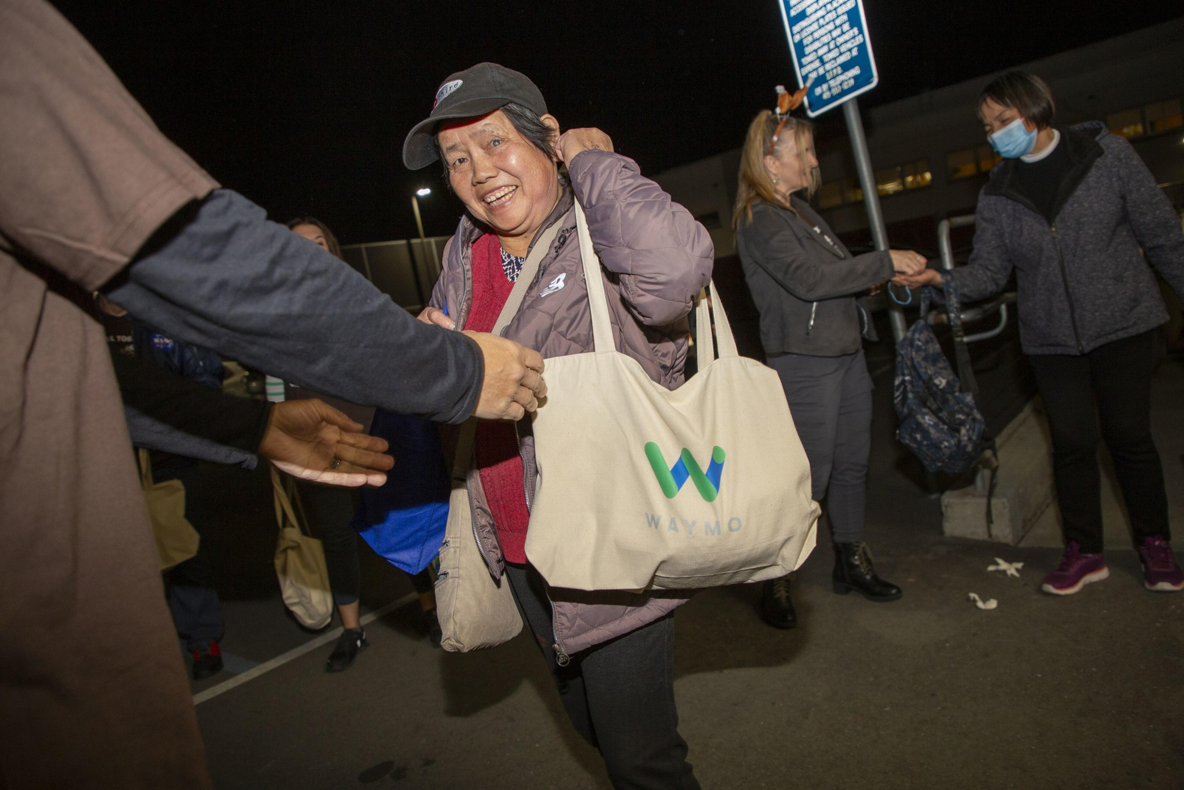 A smiling woman wearing a cap holds a Waymo tote bag among a group of people interacting at night, some of whom are exchanging handshakes.