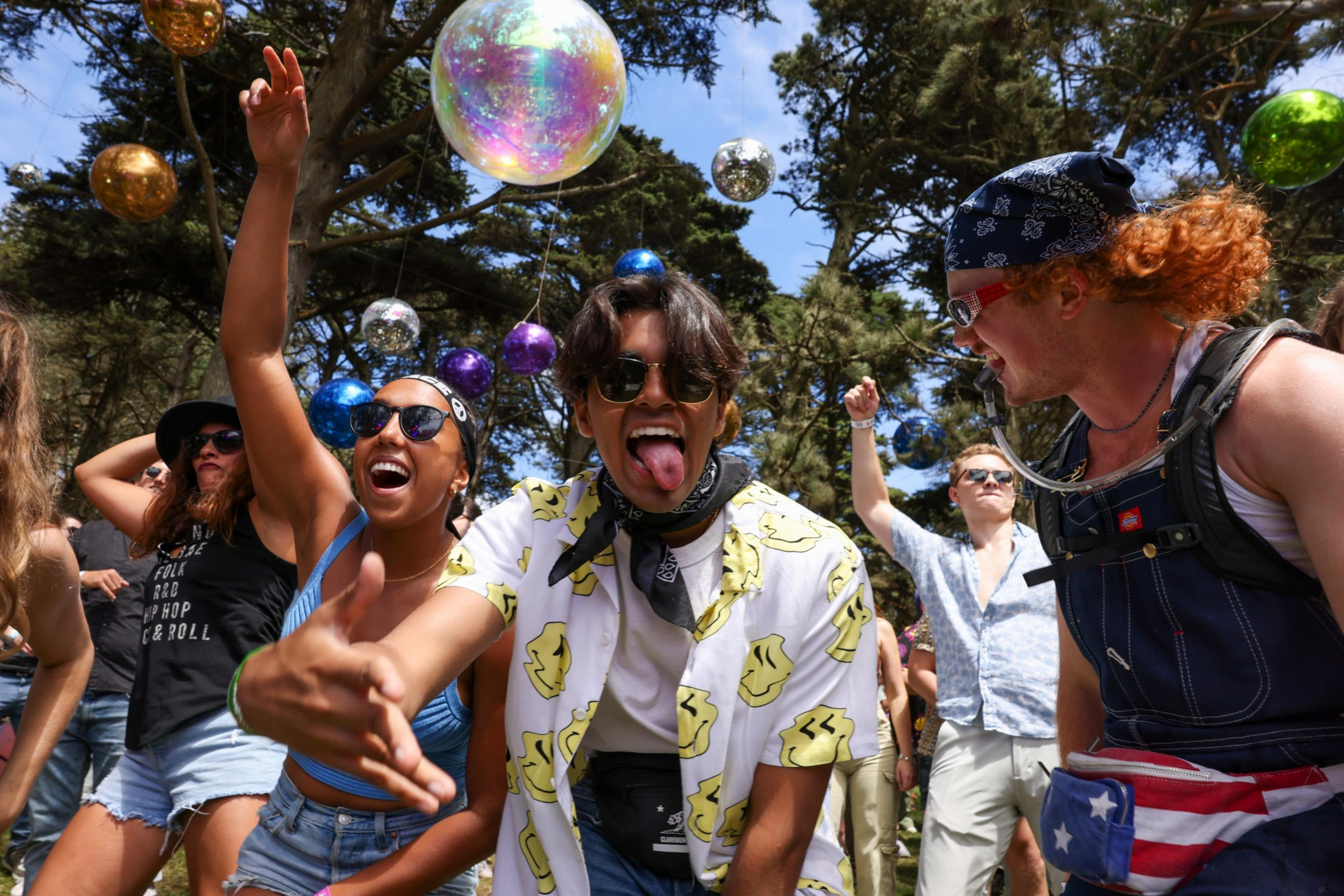A group of young people dance and smile outdoors under trees decorated with colorful hanging orbs on a sunny day.
