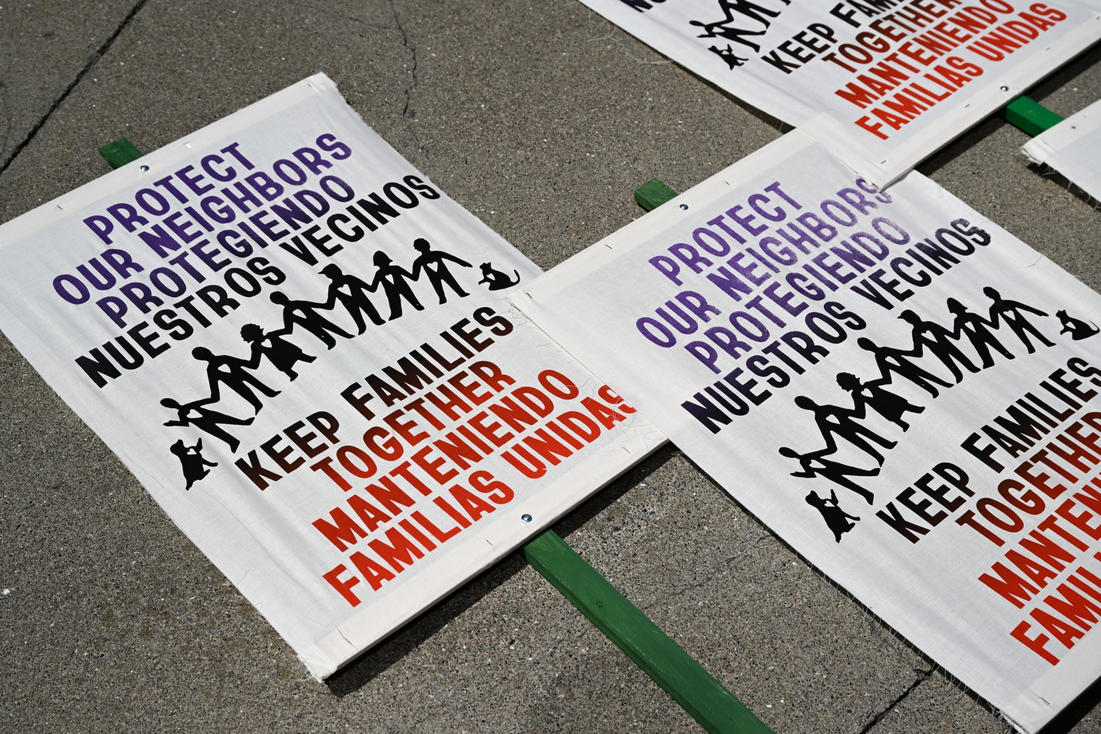 The image shows protest signs with text in English and Spanish reading, &quot;Protect Our Neighbors&quot; and &quot;Keep Families Together,&quot; placed on a concrete surface.
