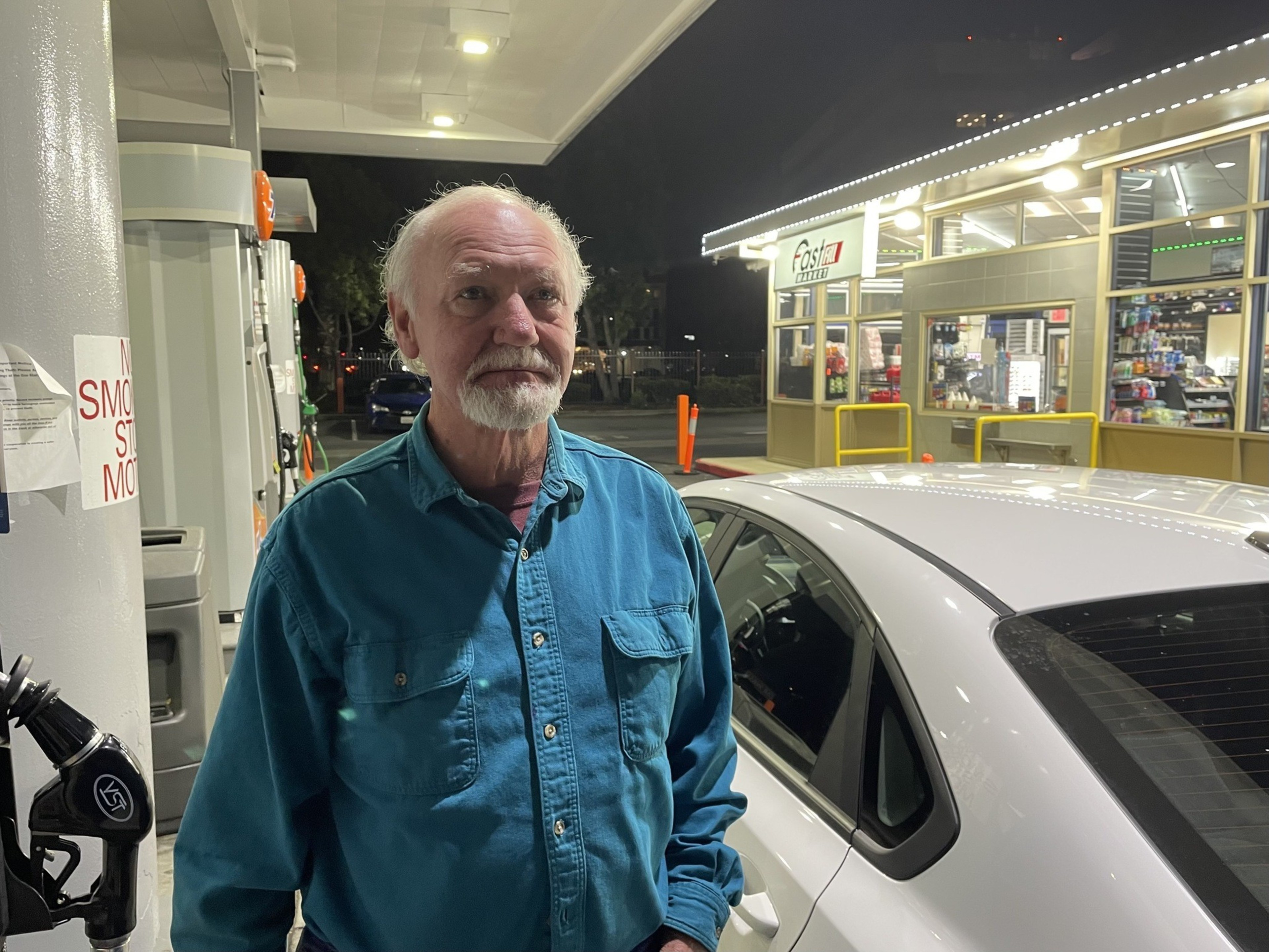 A man stands near a gas pump where his car is parked.