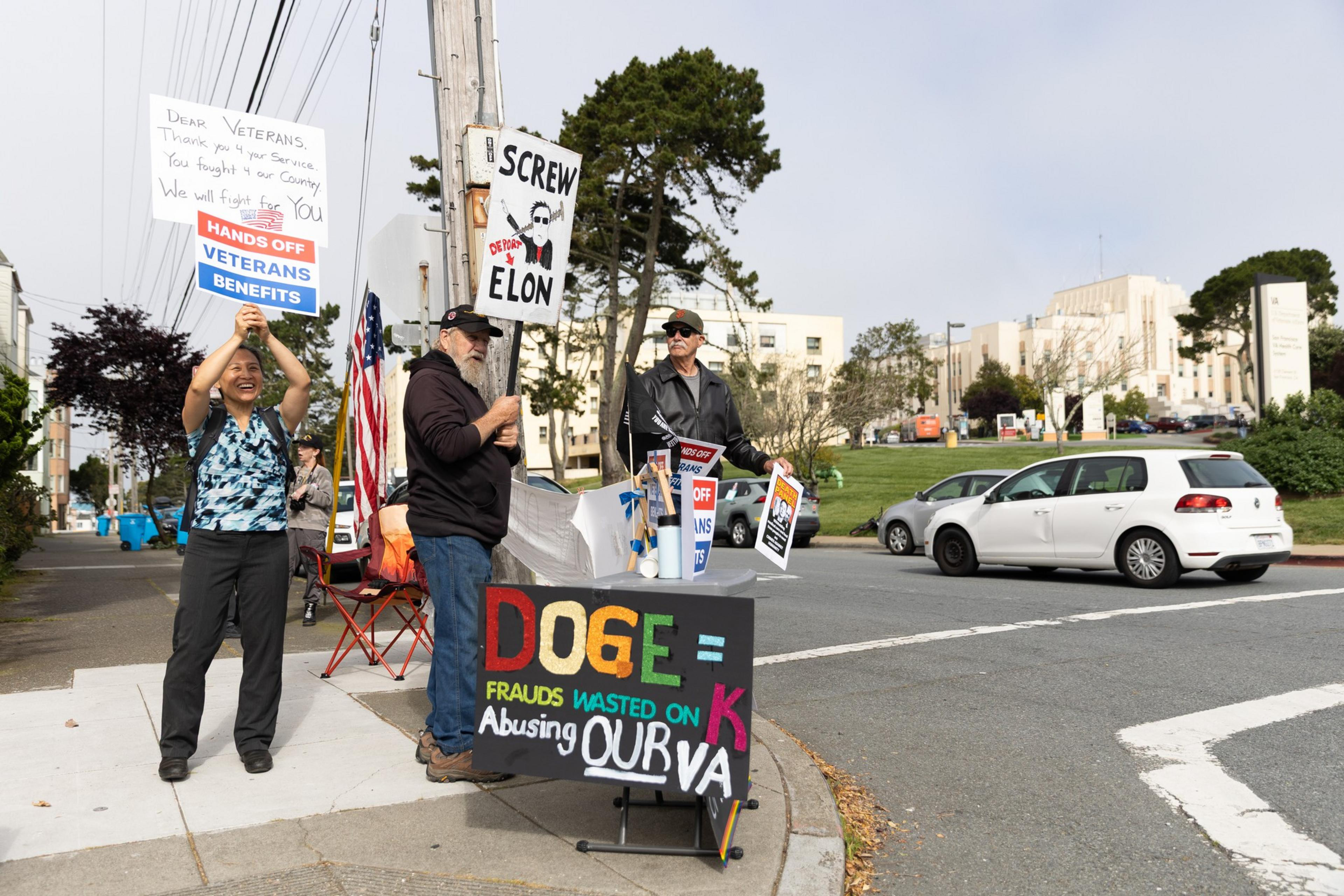 Three protesters stand on a sidewalk holding signs criticizing Elon Musk and advocating for veterans' benefits, while a car passes by on the street.