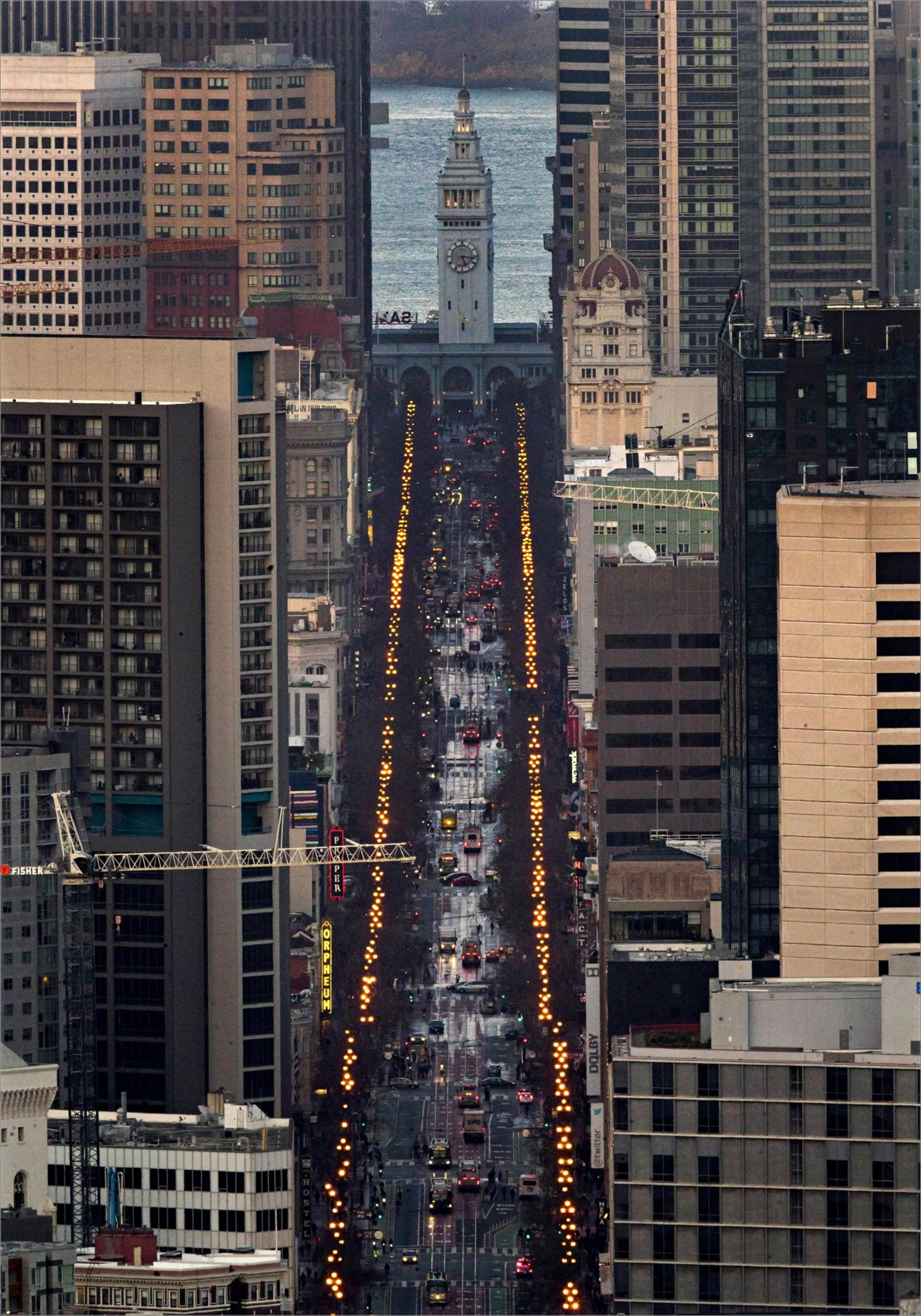A busy urban street lined with tall buildings leads to a clock tower near the waterfront, with rows of orange streetlights glowing on both sides.