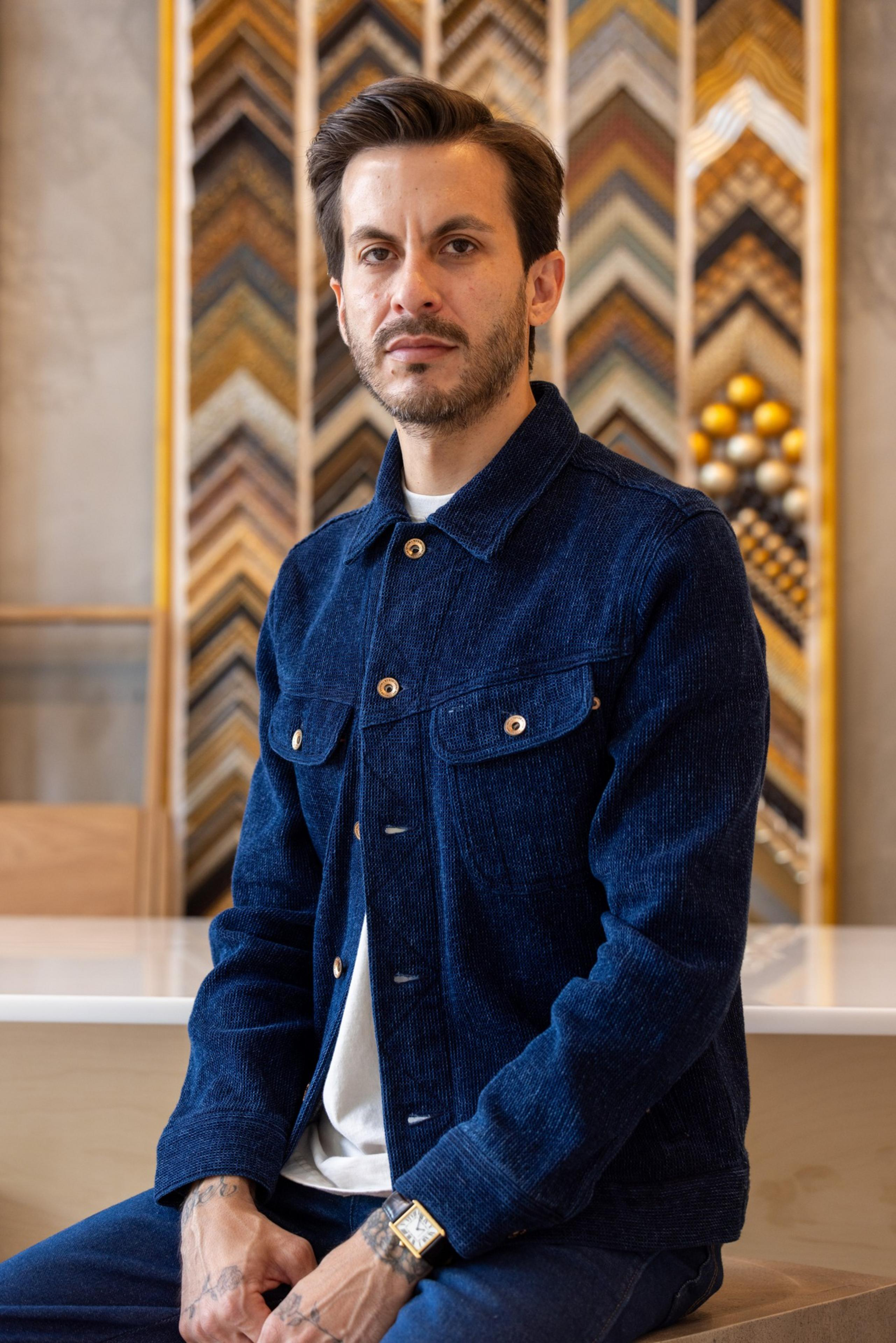 A man wearing a dark blue jacket sits in a room with a patterned background. He has short hair, a trimmed beard, and tattoos on his hands and wrist.