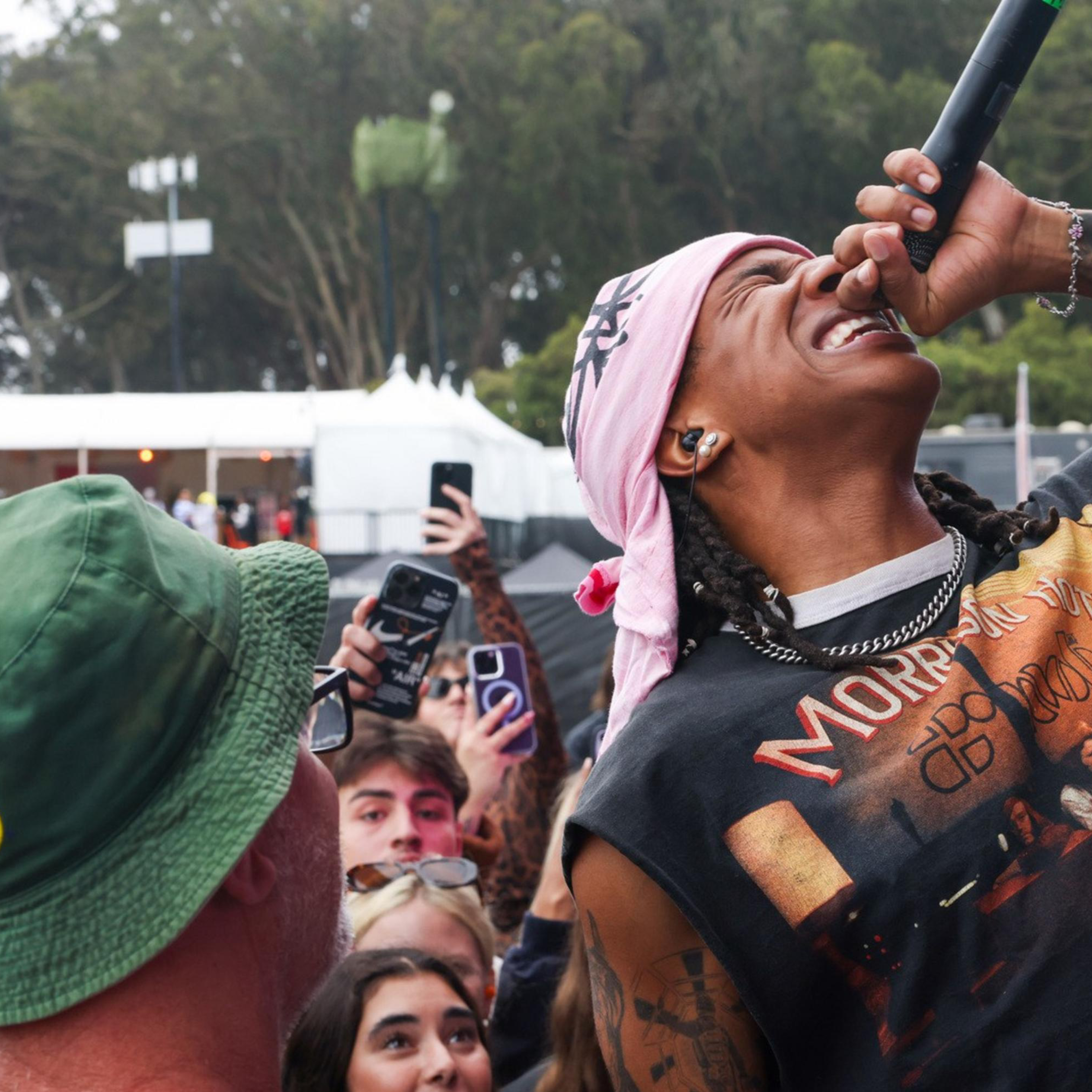 A person in a bandana sings passionately into a microphone while a crowd of fans, some recording with their phones, look on excitedly. One fan is wearing a green hat.