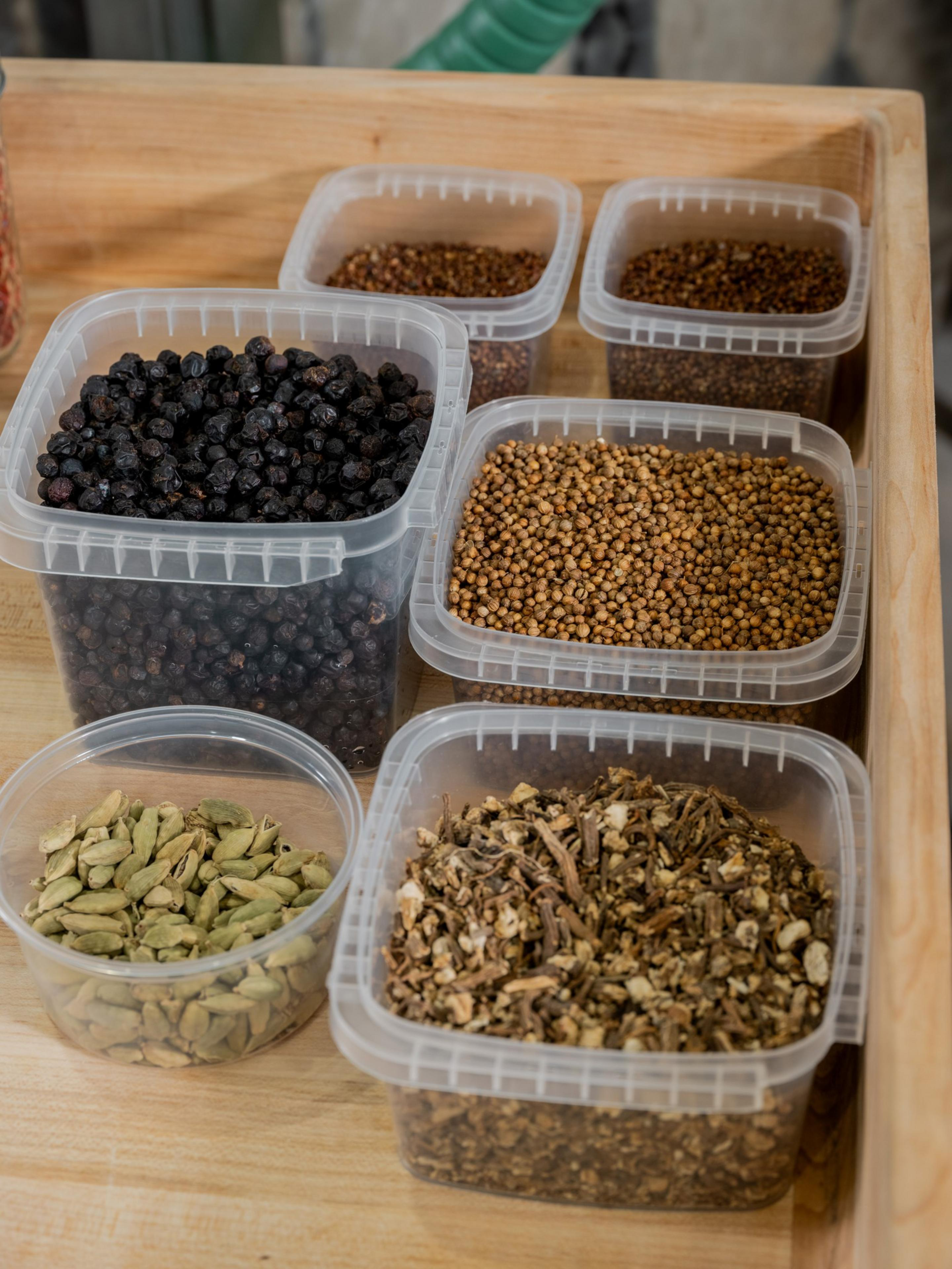 The image shows clear plastic containers filled with assorted spices, including black peppercorns, cardamom pods, coriander seeds, and dried root pieces on a wooden surface.