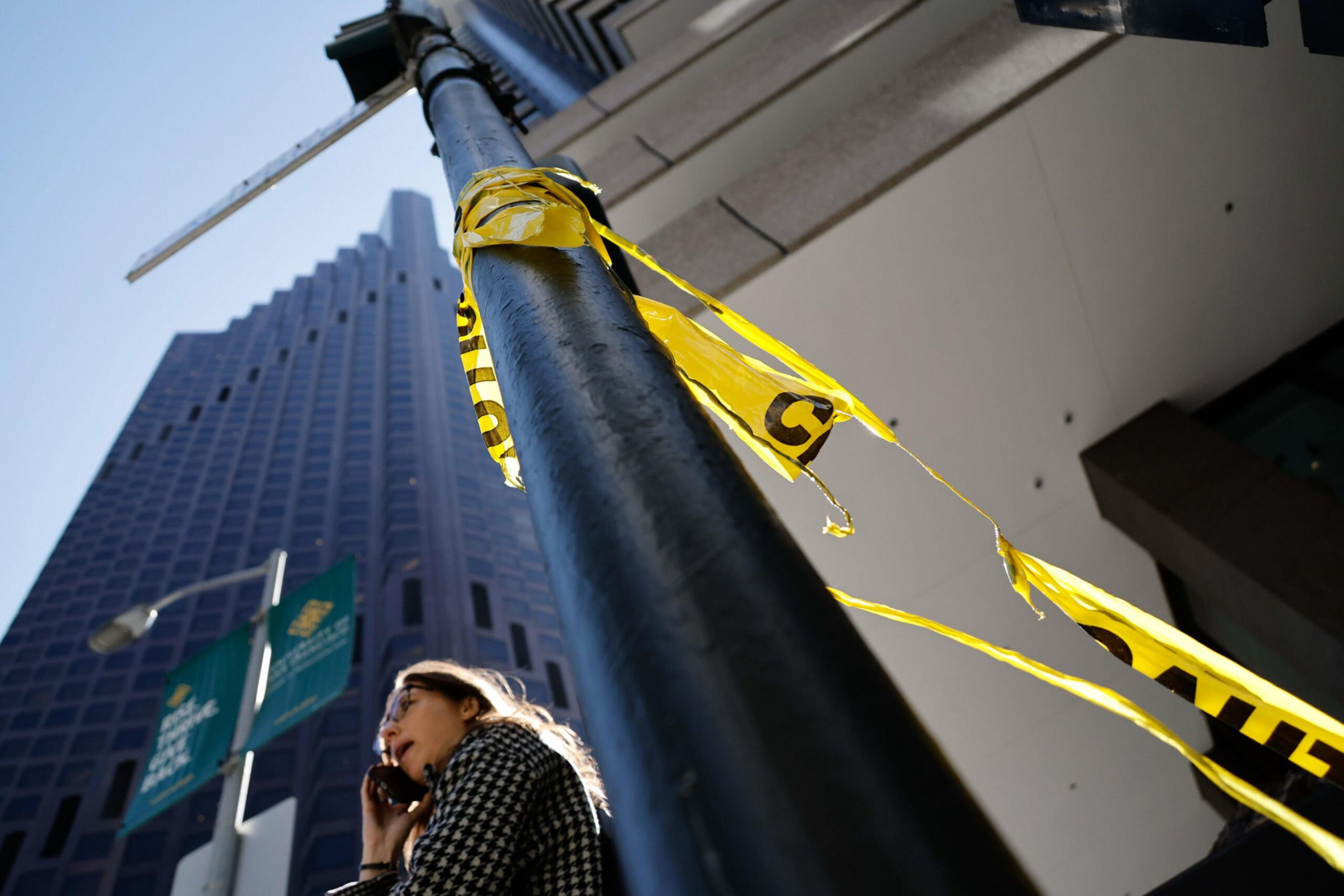 A woman in a checkered coat talks on a phone near a tall black pole wrapped with torn yellow caution tape, with skyscrapers and banners in the background.