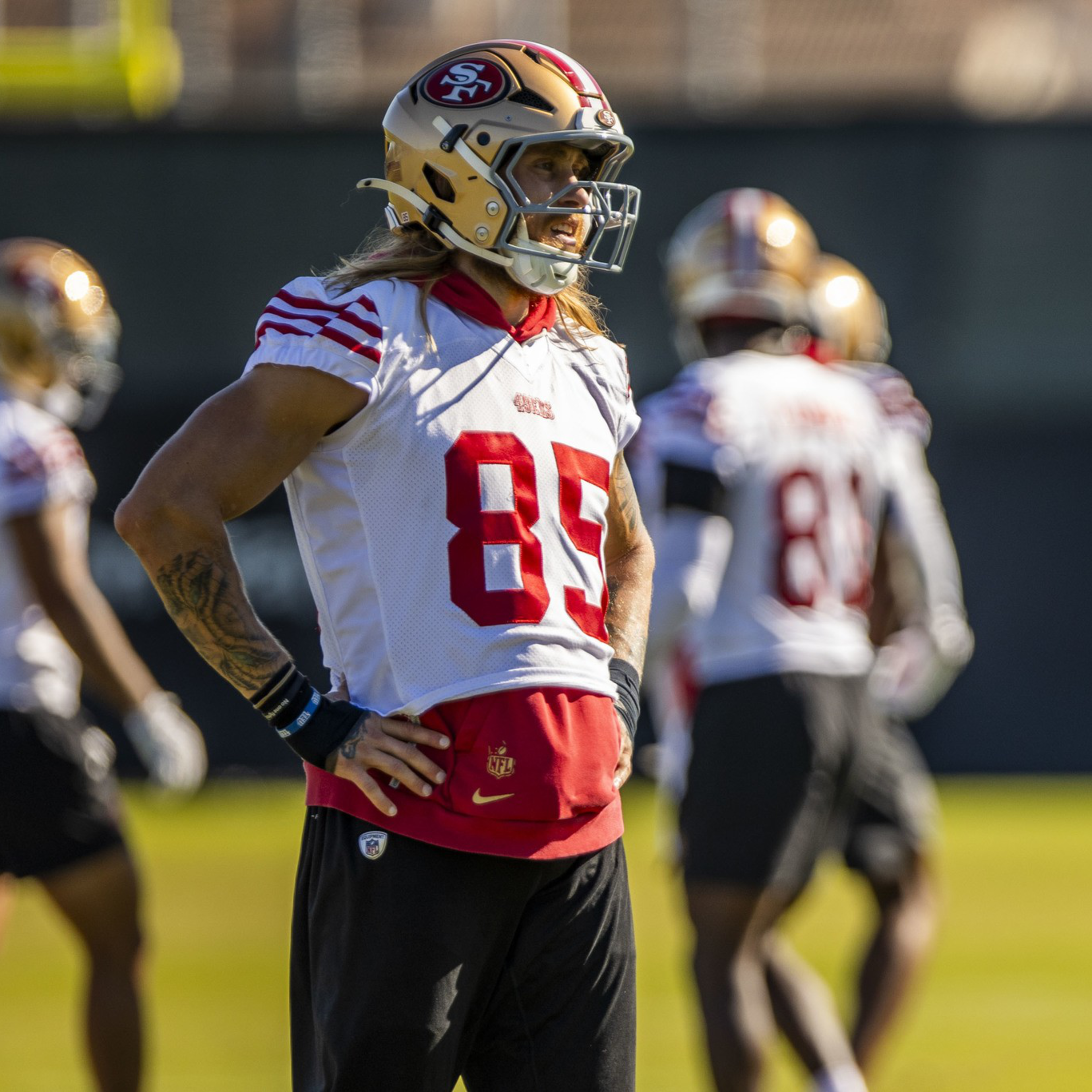 A football player wearing a white and red San Francisco 49ers jersey number 85 stands with hands on hips while teammates practice in the background.