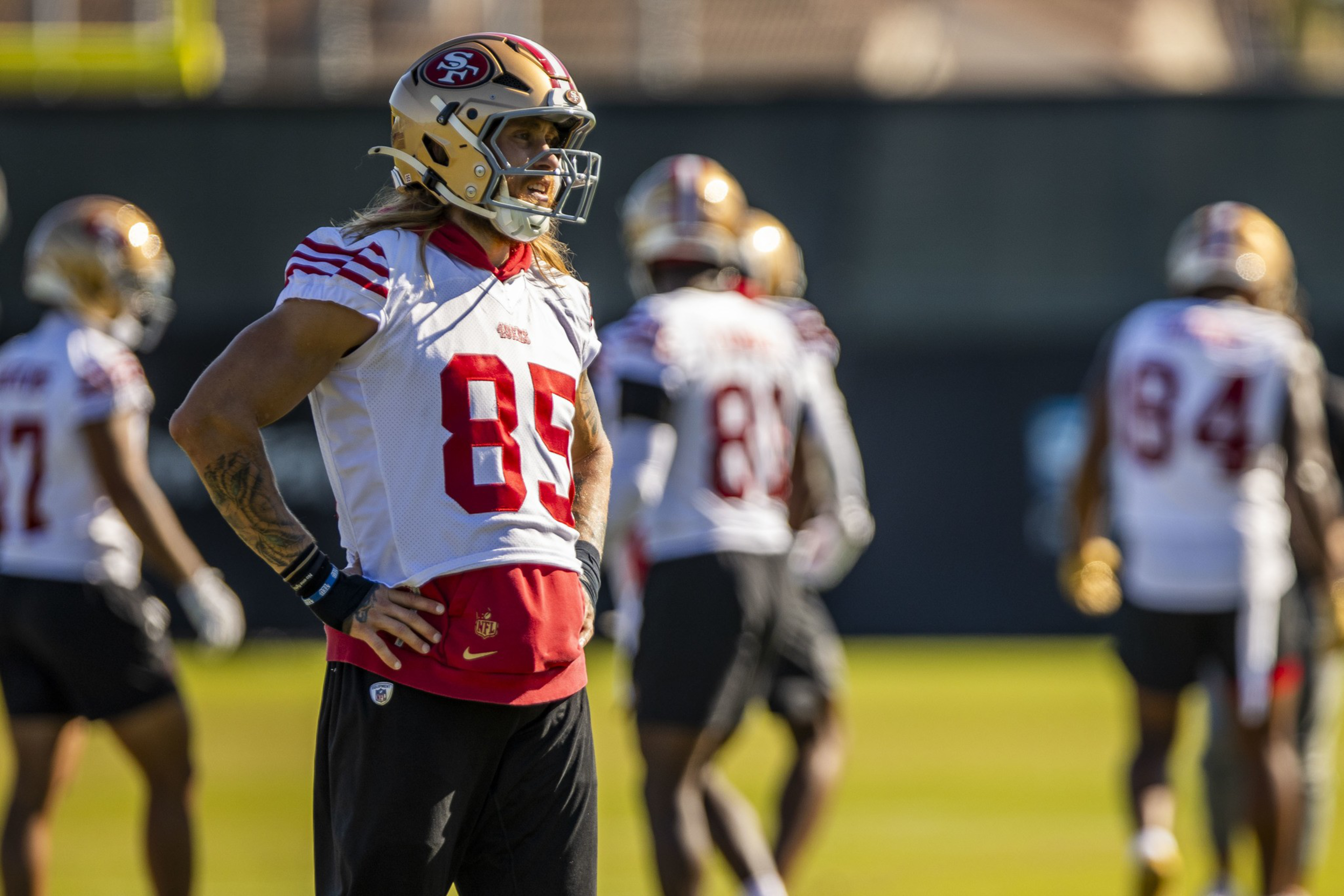 A football player wearing a white and red San Francisco 49ers jersey number 85 stands with hands on hips while teammates practice in the background.