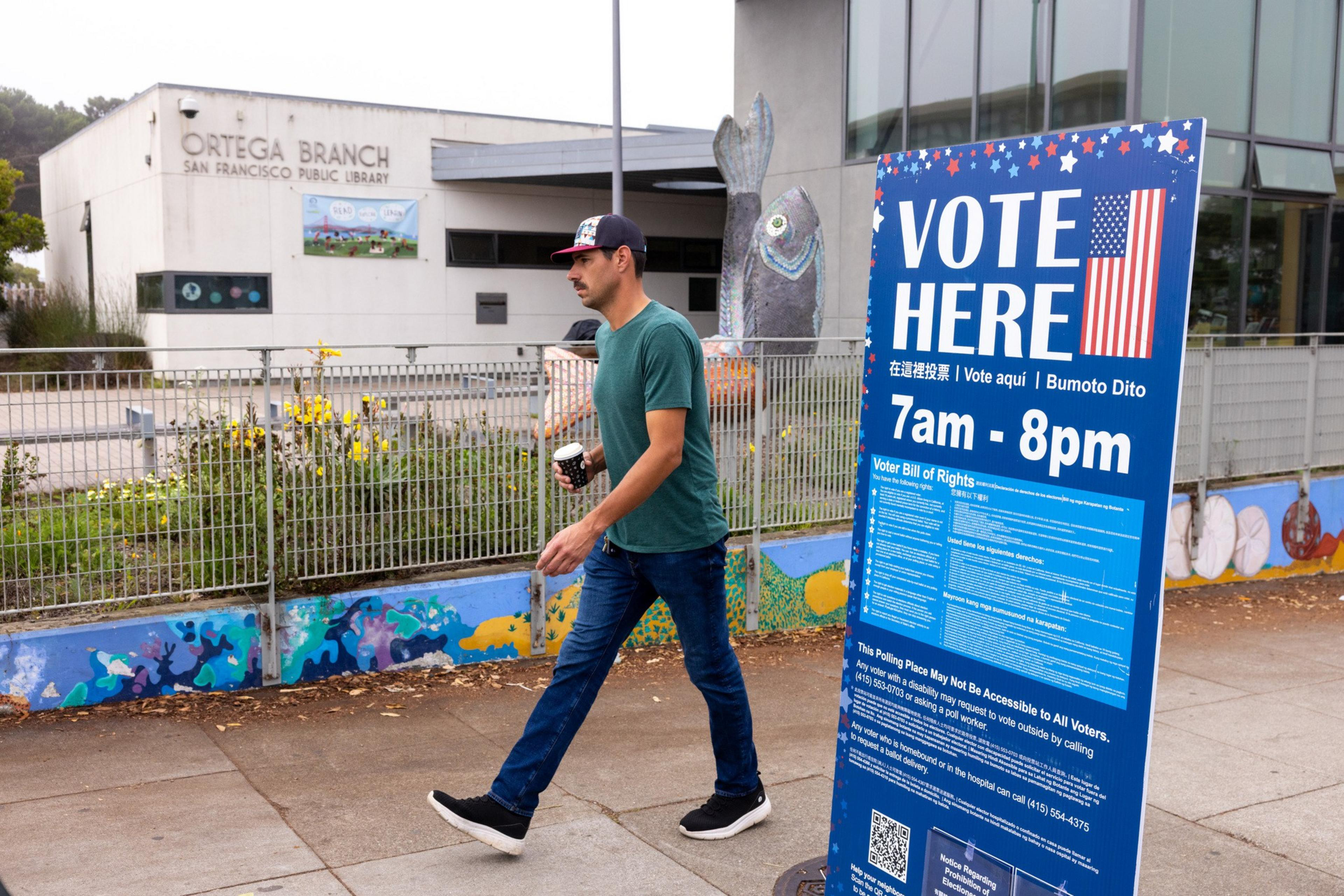 A man walks past a colorful fence outside Ortega Branch San Francisco Public Library near a large blue “Vote Here” sign displaying voting hours from 7 am to 8 pm.
