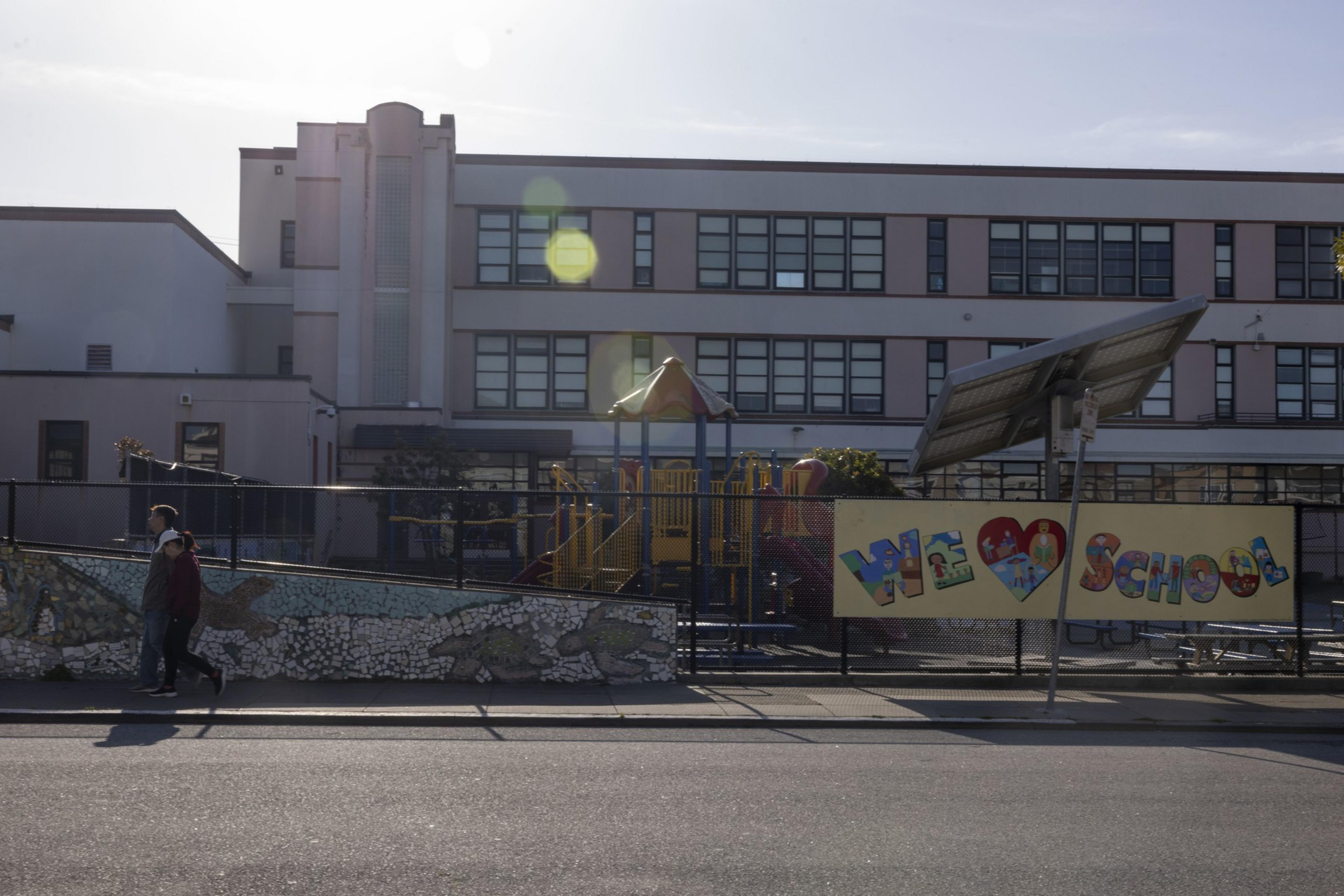 A school building with large windows is in the background. A colorful playground is behind a fence with a &quot;We Love School&quot; banner. Two people walk past.