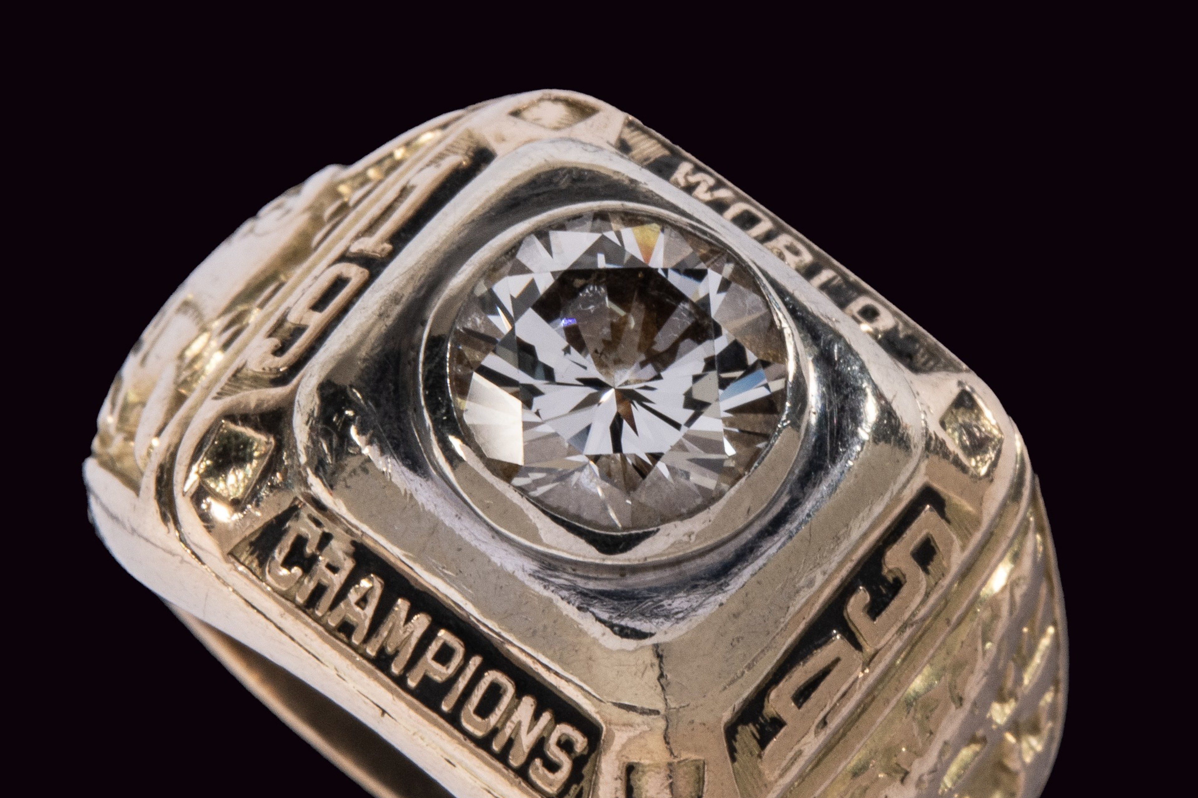 The image shows a gold championship ring with a large central diamond. It has the words "World Champions" engraved around the diamond.