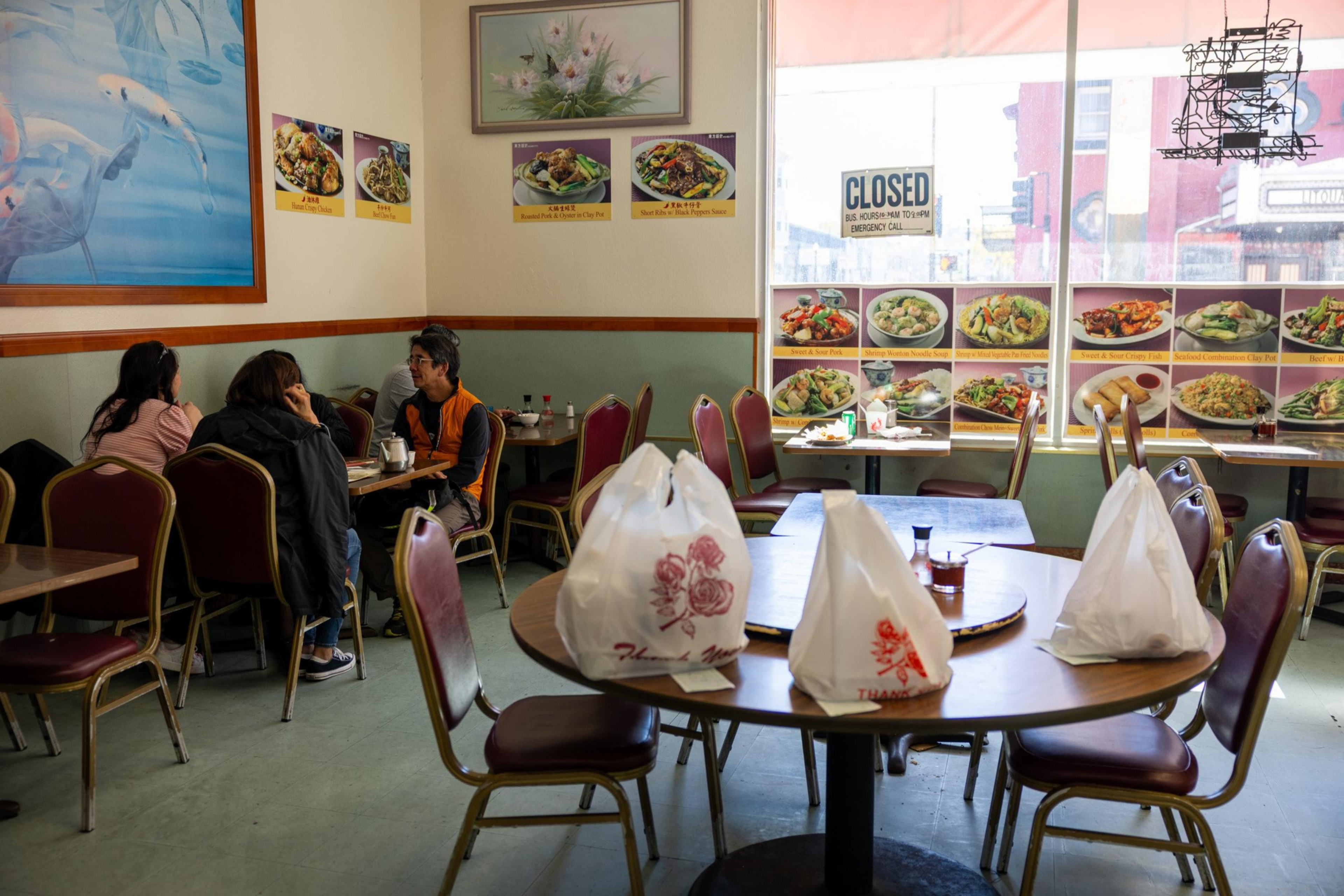 Three people sit and talk at a table in a small restaurant with food pictures on the walls and many takeout bags on a nearby table.