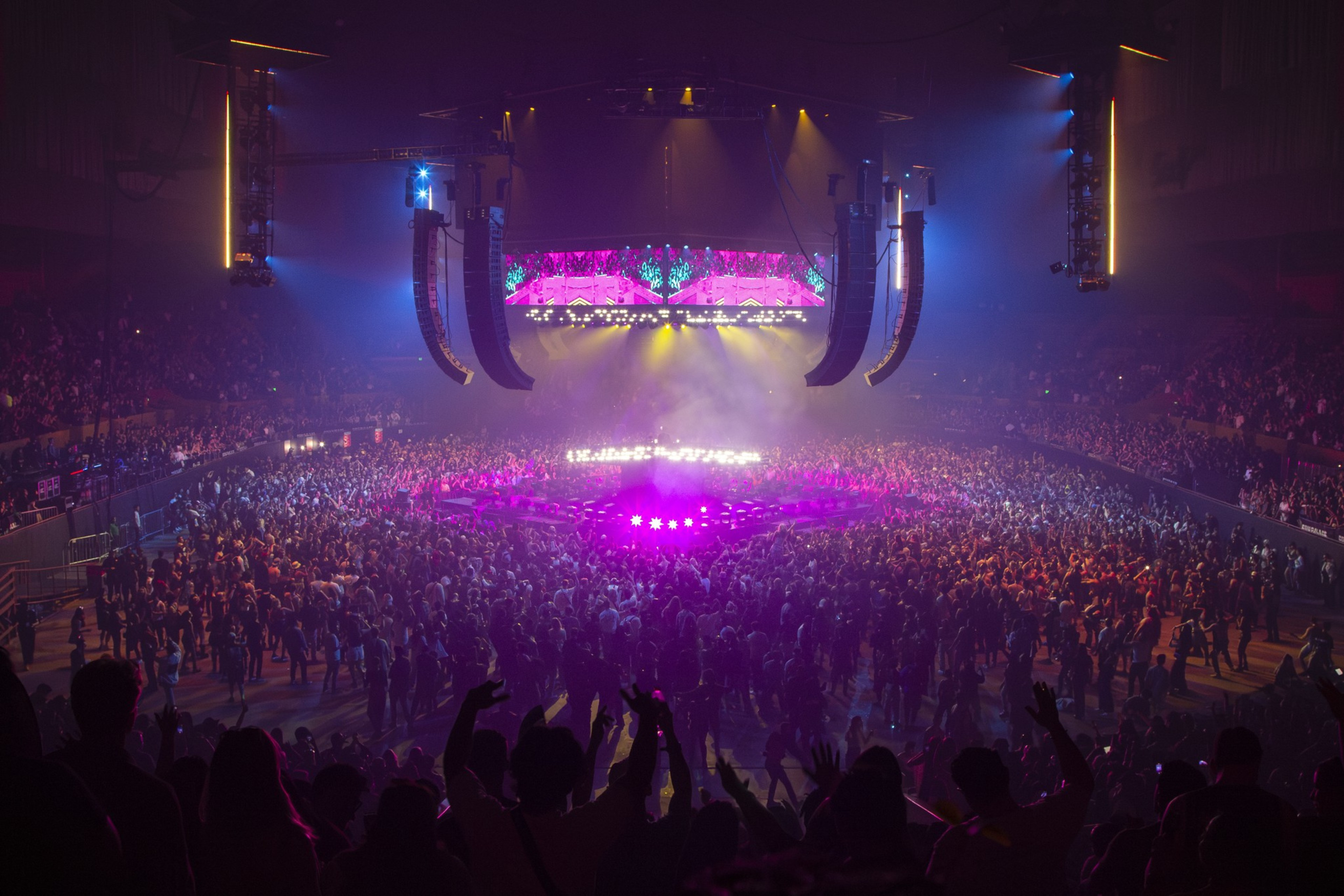 A crowded concert venue features a brightly lit, circular stage surrounded by an audience. Colorful lights and large speakers create an energetic atmosphere.
