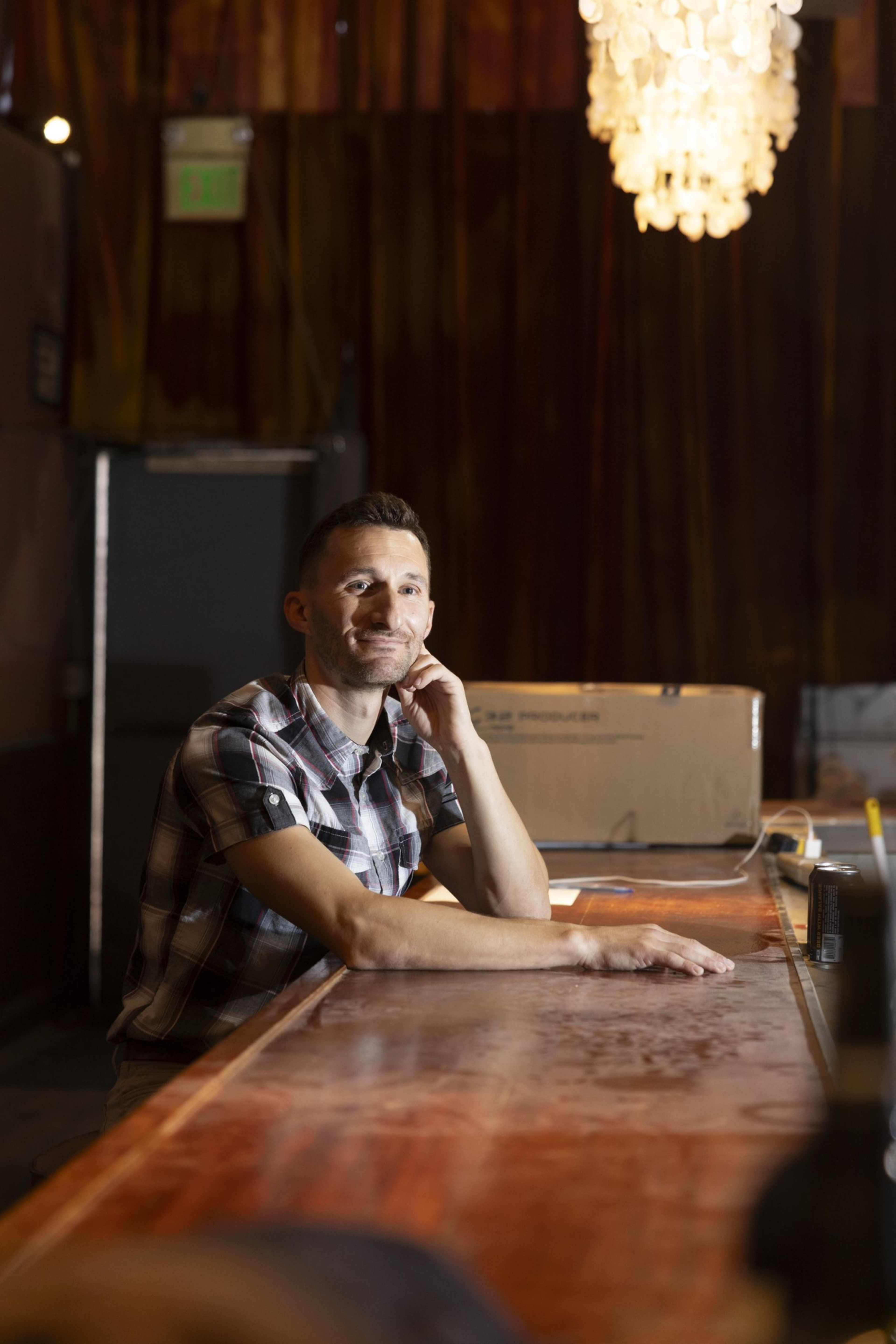 A man in a plaid shirt sits at a wooden bar, resting his chin on his hand, with a chandelier hanging above and a cardboard box in the background.