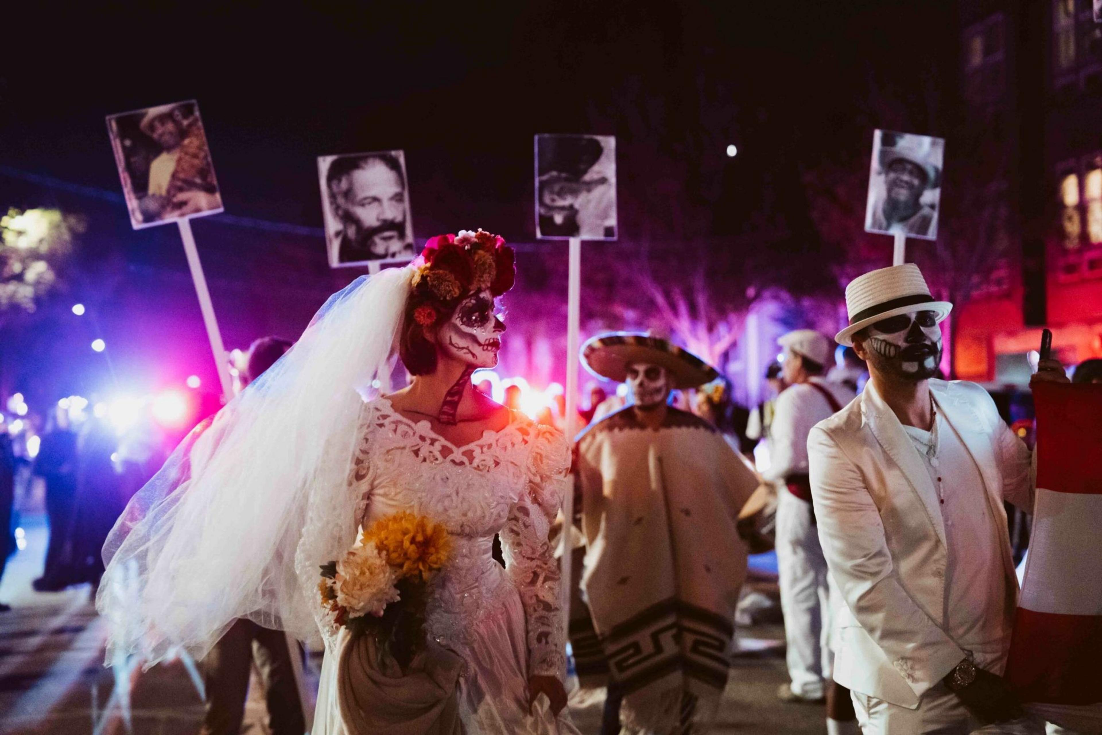 People dressed in Día de los Muertos costumes, including a bride with a flower crown and face paint, march holding black-and-white photos on sticks.