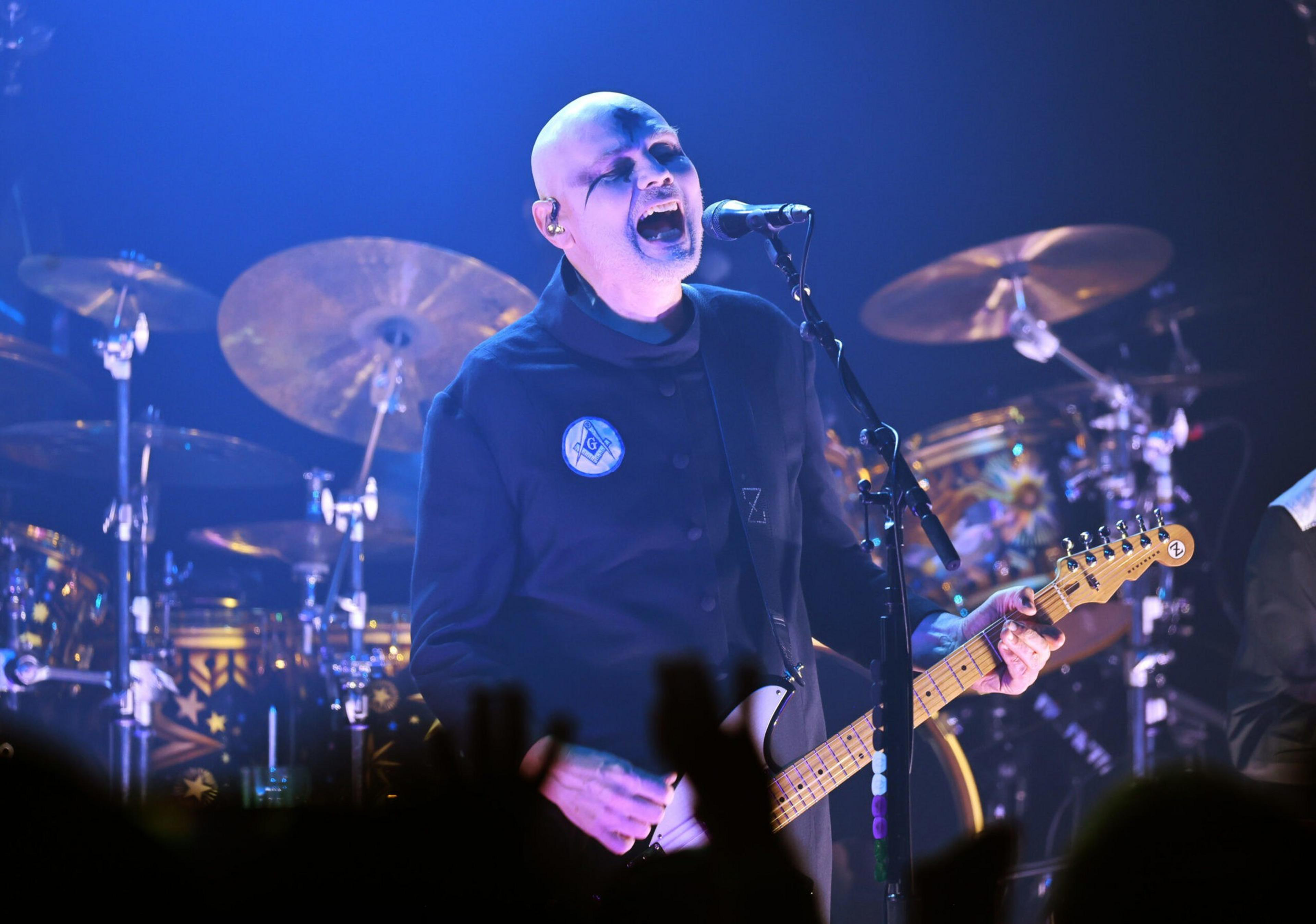 A bald musician passionately sings and plays electric guitar on stage, with drums and cymbals visible behind him and audience hands raised in front.