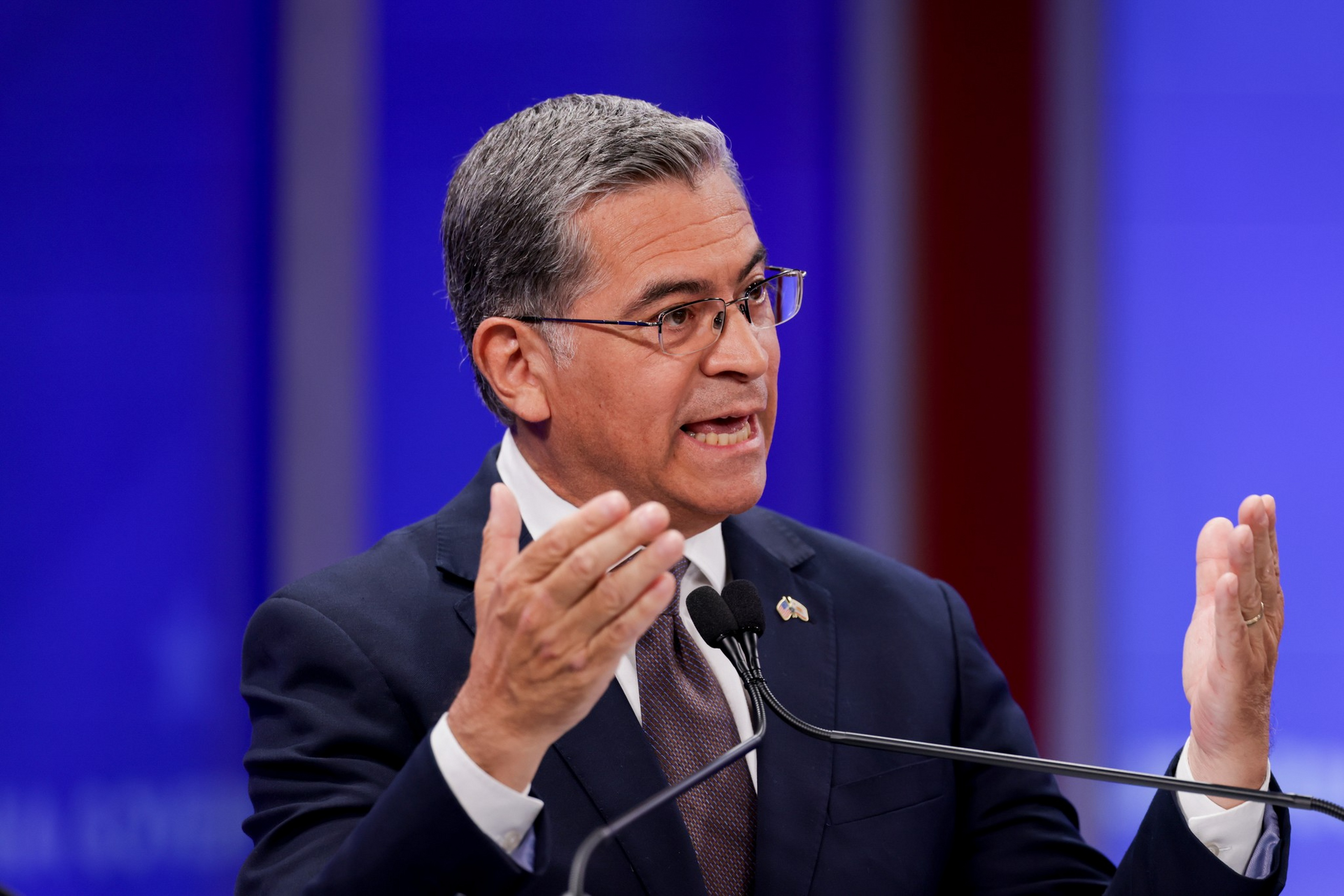 A man in a dark suit and glasses speaks passionately into a microphone, gesturing with both hands, against a blue and red background.