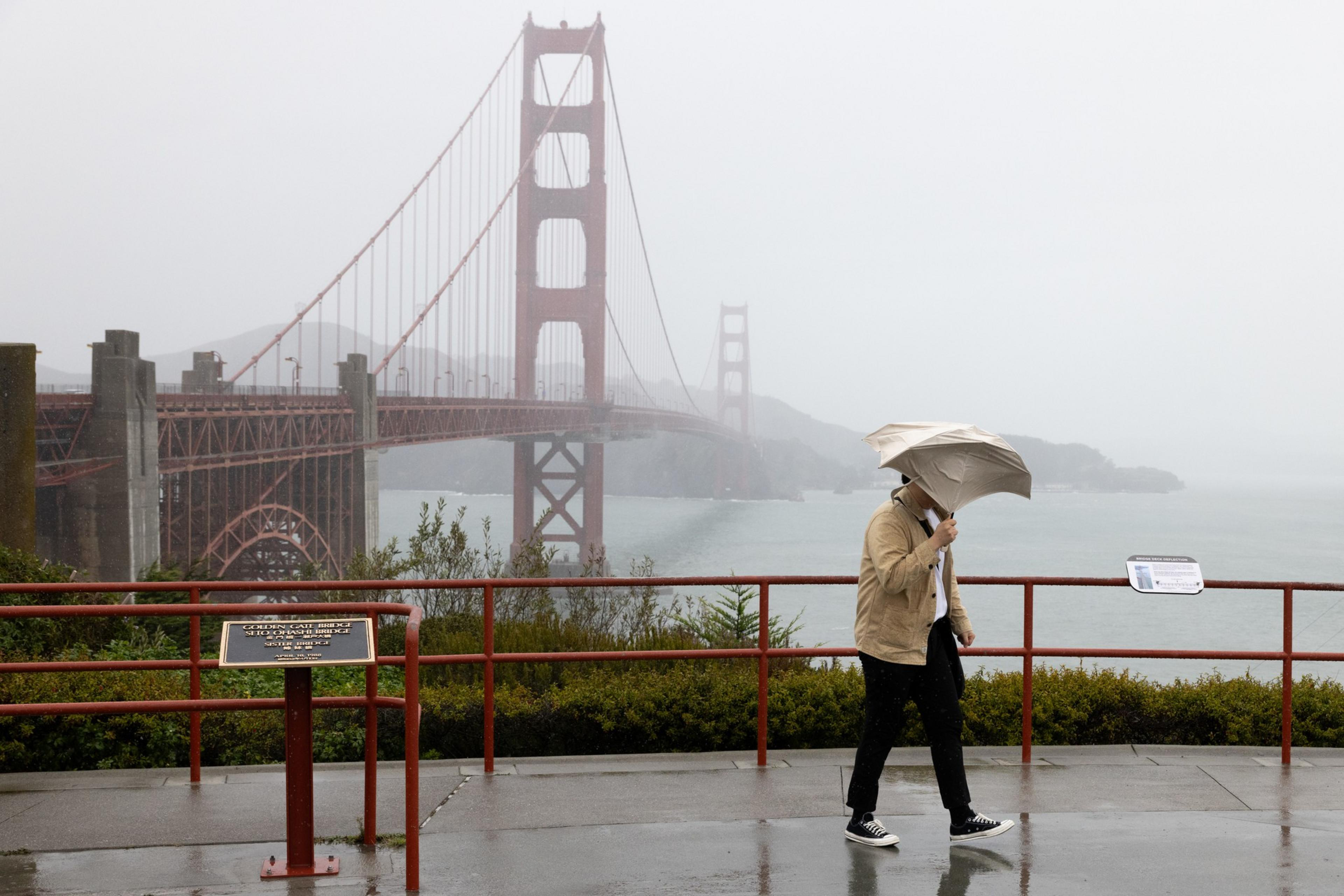 A person with an umbrella walks in the rain near the Golden Gate Bridge, which is partially obscured by mist. The scene is set on a gray, overcast day.