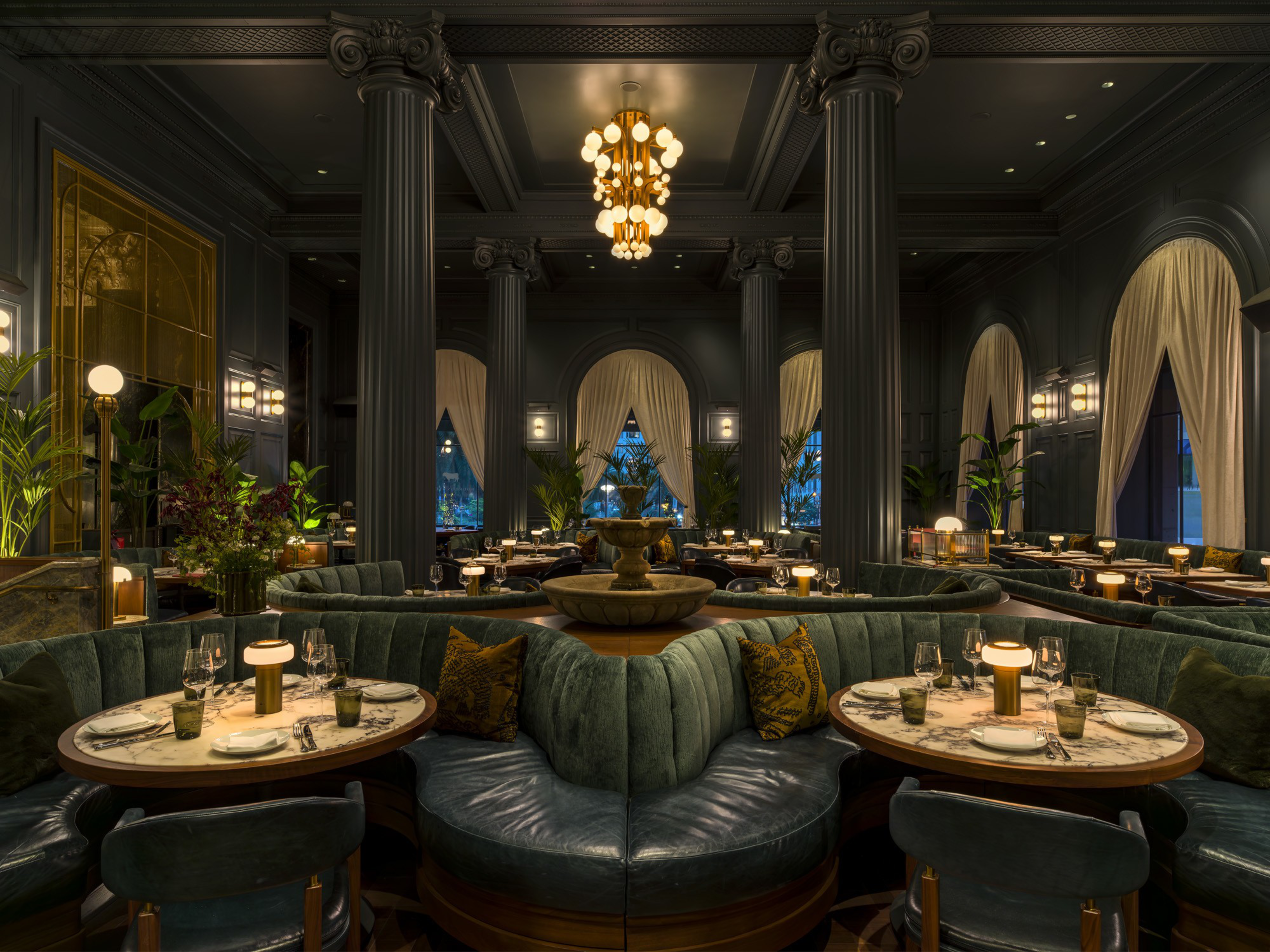 A dimly lit upscale restaurant with plush green seating, marble tables, tall pillars, large draped windows, plants, and a central ornate chandelier.