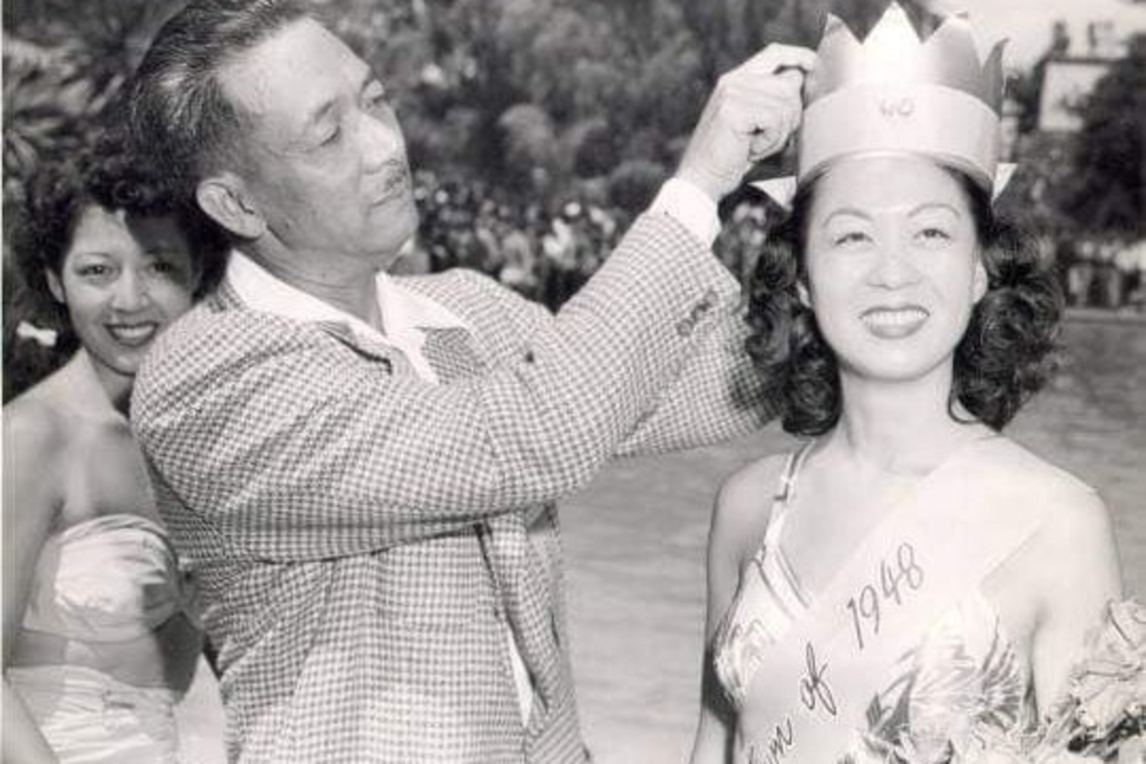 A man places a paper crown on a smiling woman wearing a "Queen of 1948" sash, with another smiling woman in a strapless dress in the background outdoors.