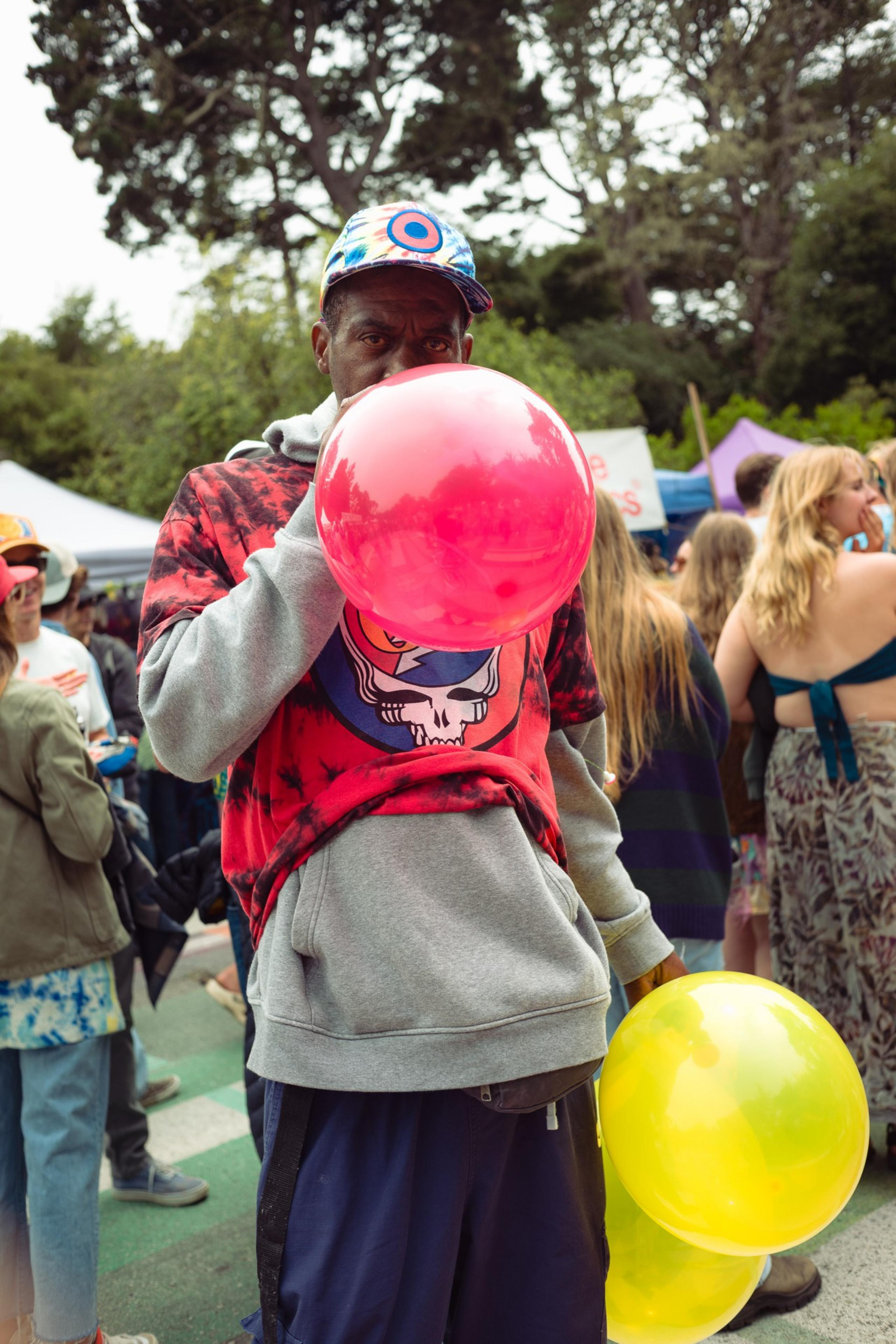 A man wearing a colorful cap and a red graphic shirt over a gray hoodie is inflating a red balloon while holding two yellow balloons at an outdoor event.