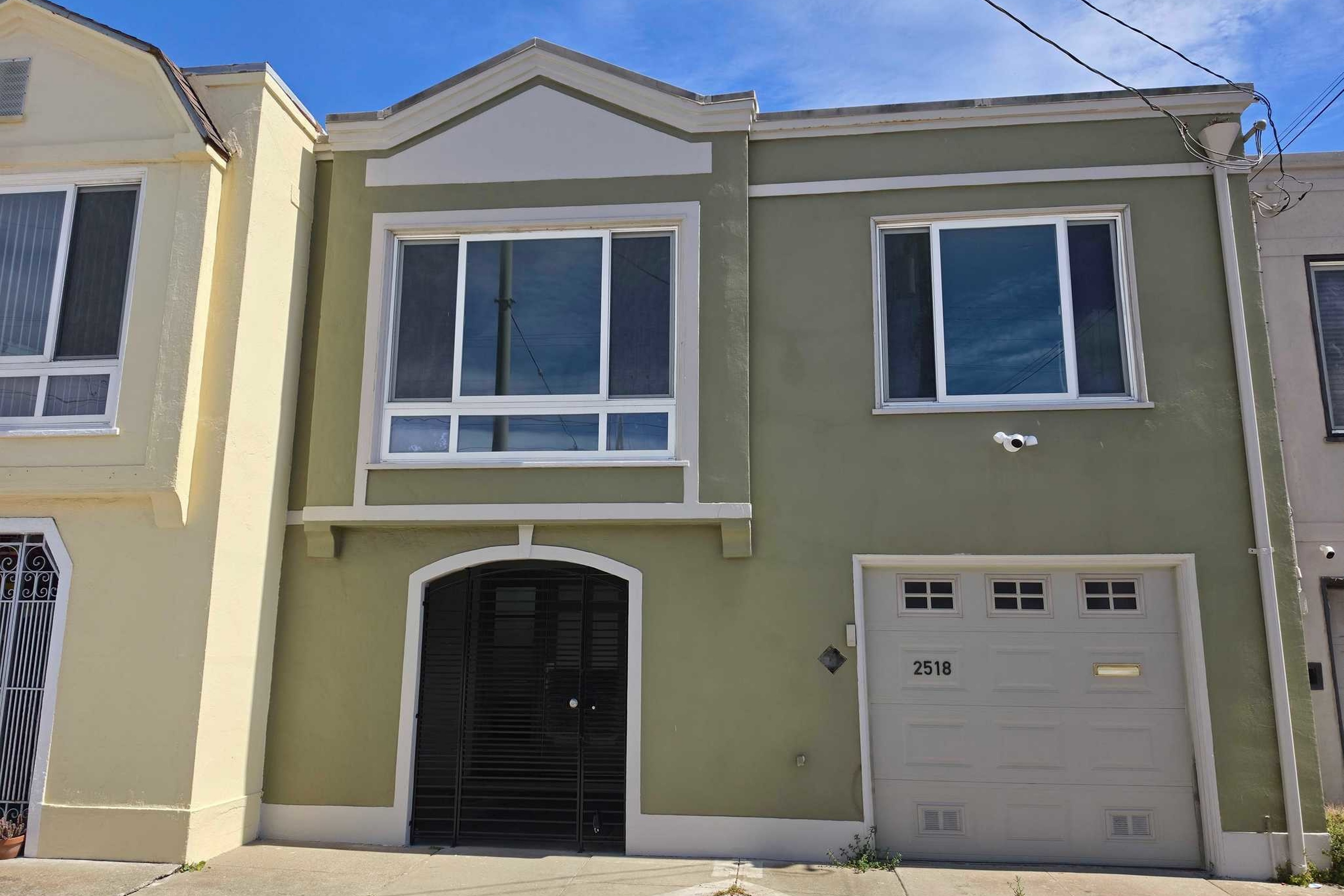A two-story olive-green house with white trim, featuring a large window above a black arched door and a garage door numbered 2518 to the right.