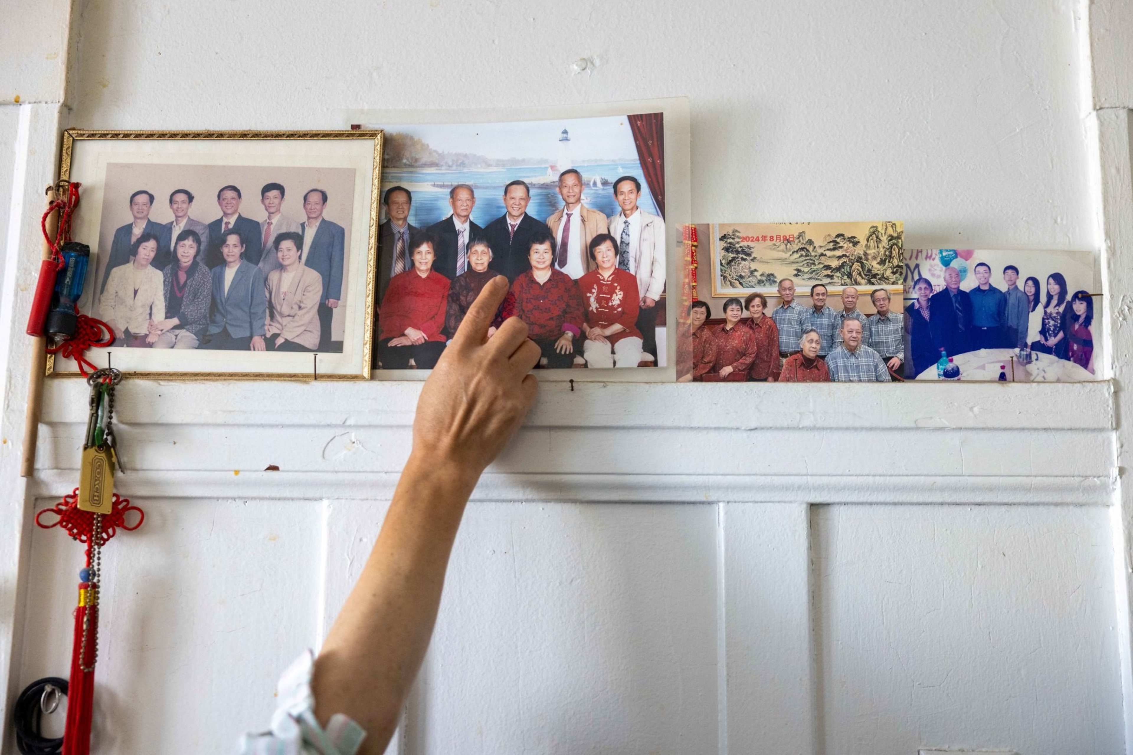 A hand points to a family photo on a wall with several framed and unframed group pictures of people arranged in a row.