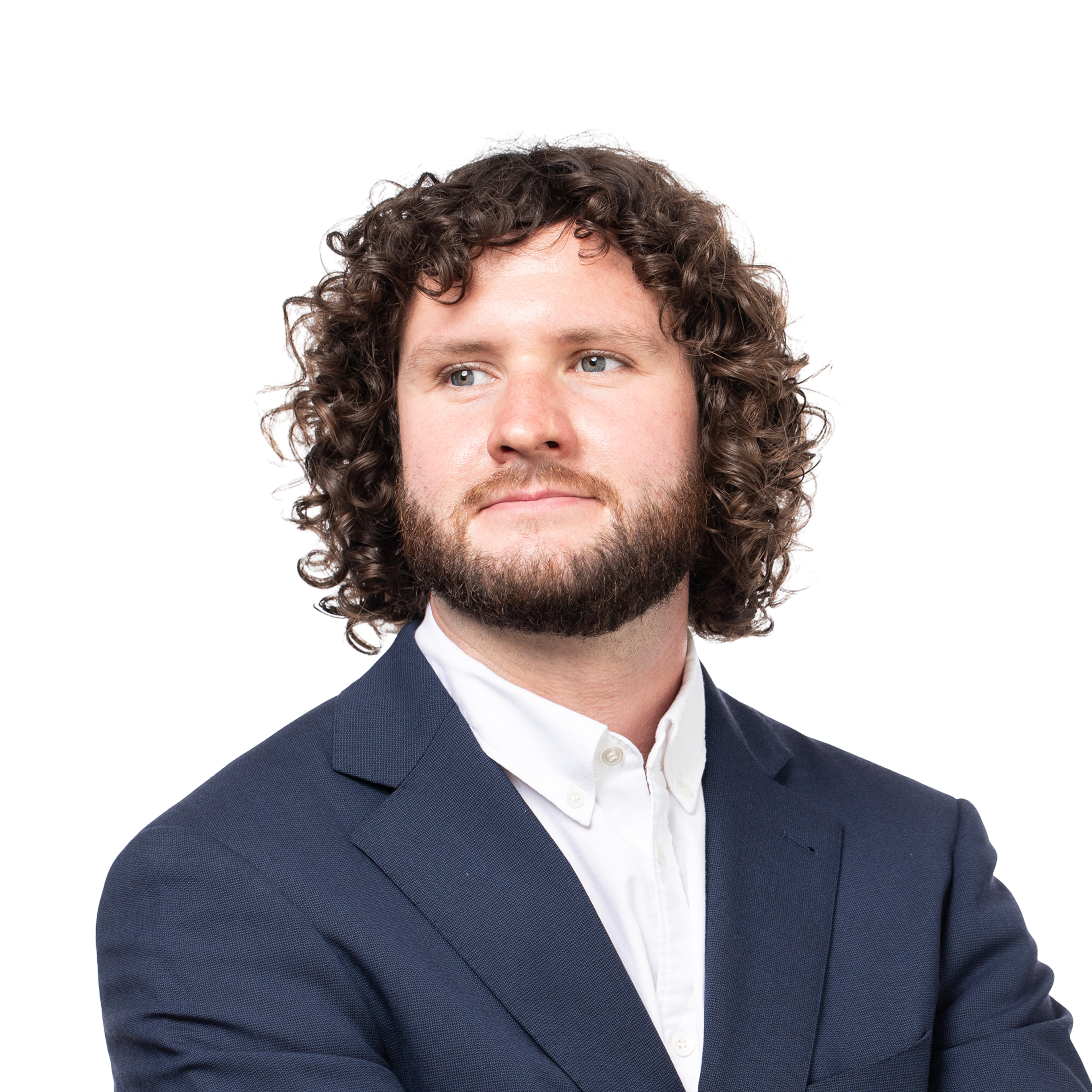 A man with curly brown hair and a beard wears a navy blue suit jacket over a white button-up shirt, looking slightly to his right with a calm expression.
