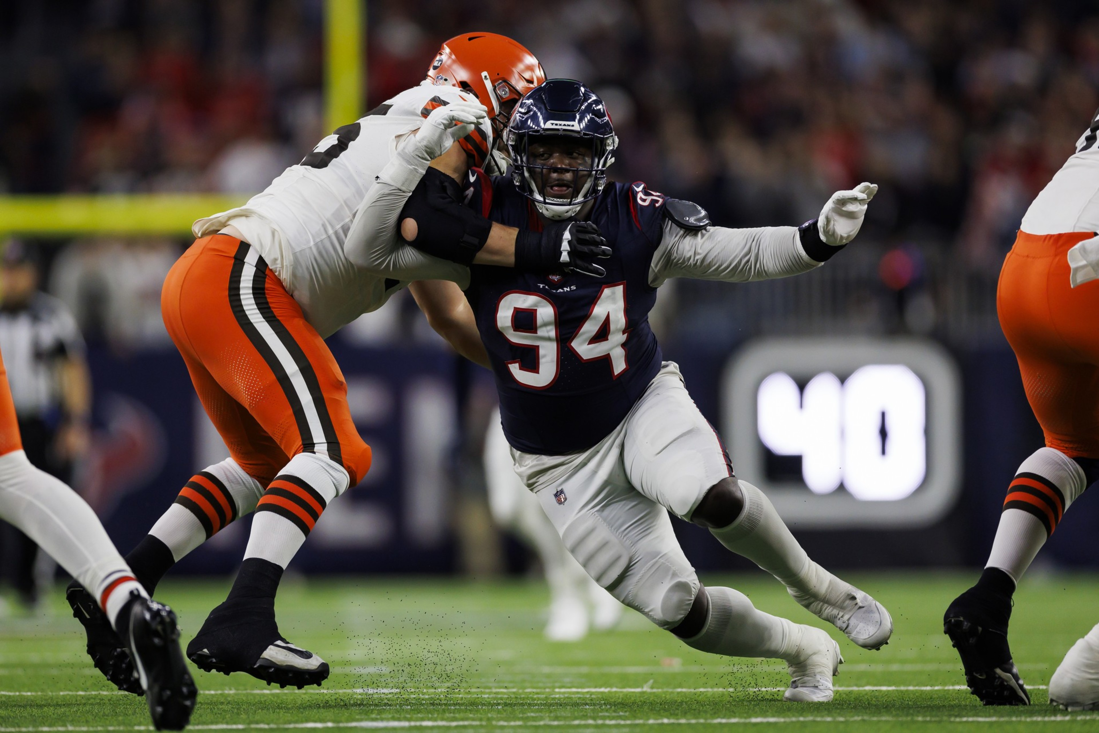 A football player in a navy uniform is tackling an opponent in a white and orange uniform on the field, with both in action, surrounded by blurred spectators.