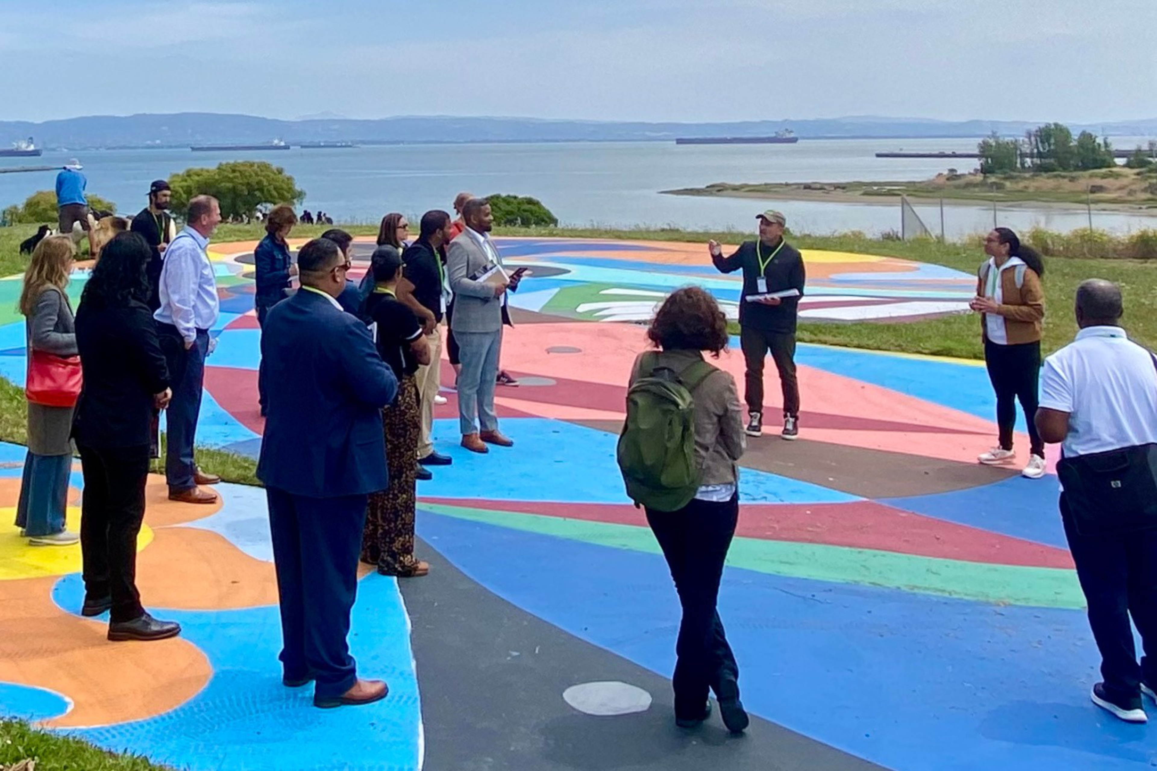 A group of people listens to a speaker outdoors on a colorful painted surface by the water, with trees and ships visible in the background.