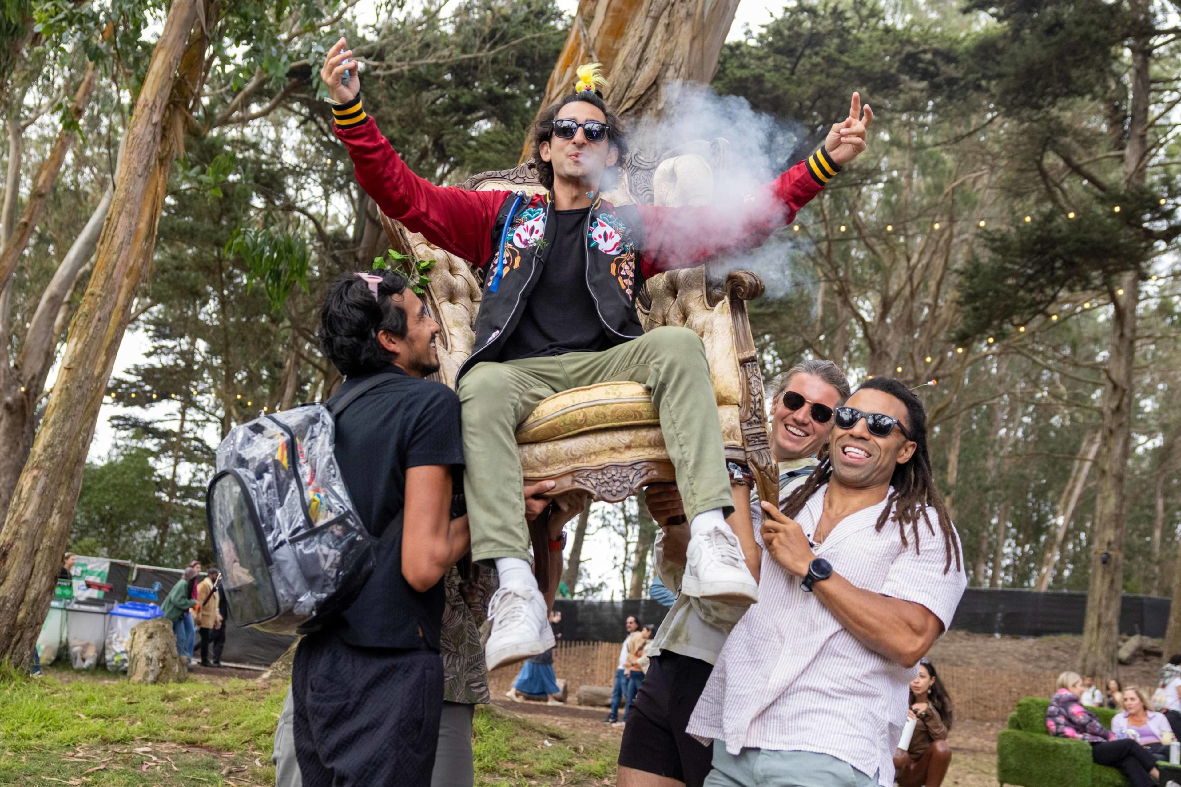 A man wearing sunglasses and a colorful jacket blows smoke while being lifted on a throne-like chair by three smiling friends outdoors among trees.