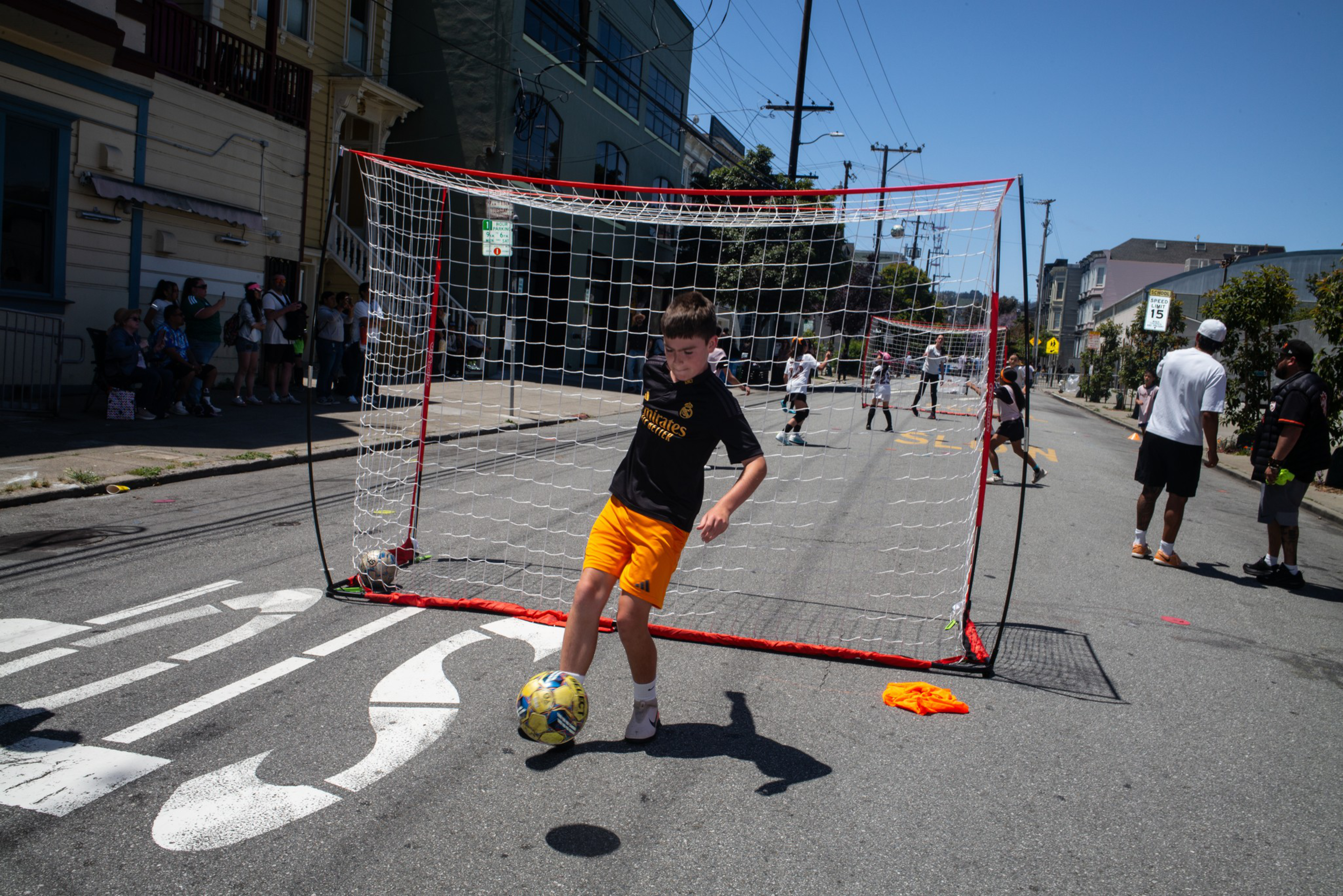 A boy in orange shorts and a black shirt controls a soccer ball near a goal set up on a city street with people watching and playing in the background.