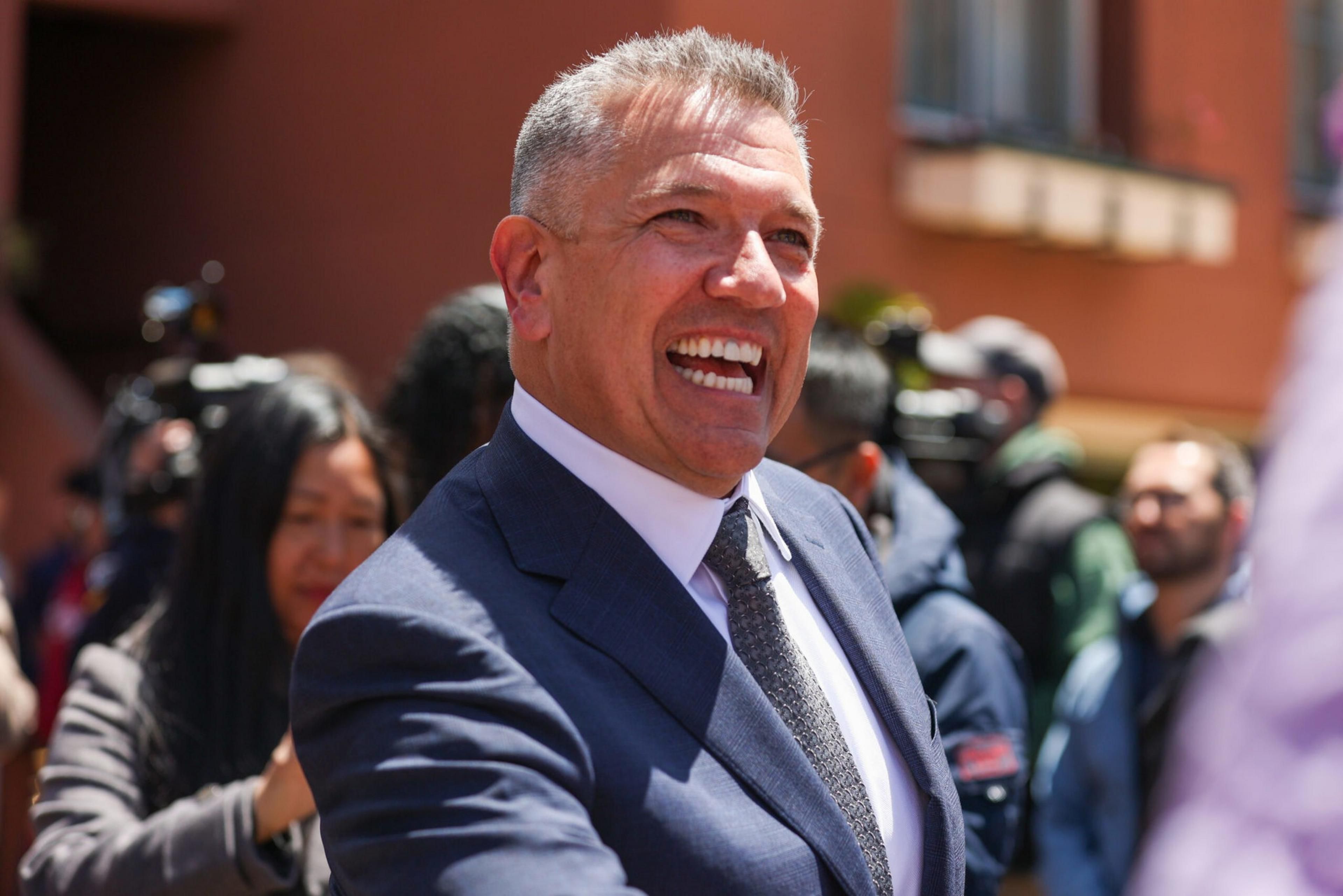 A smiling man in a dark suit and tie is shaking hands outdoors, with people and cameras blurred in the background.