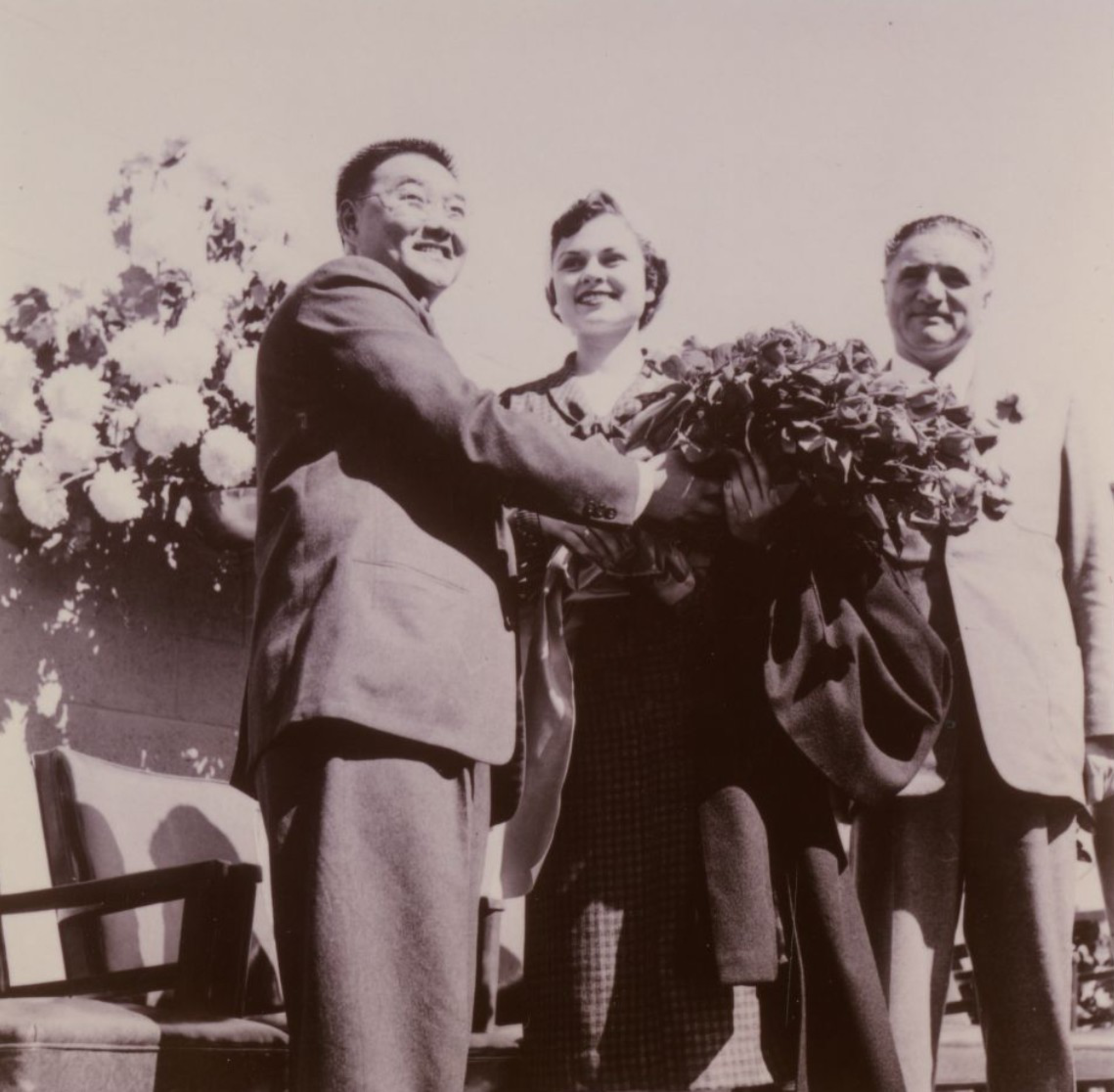 A man presents a large bouquet of flowers to a smiling woman while another man stands beside them, all dressed formally.