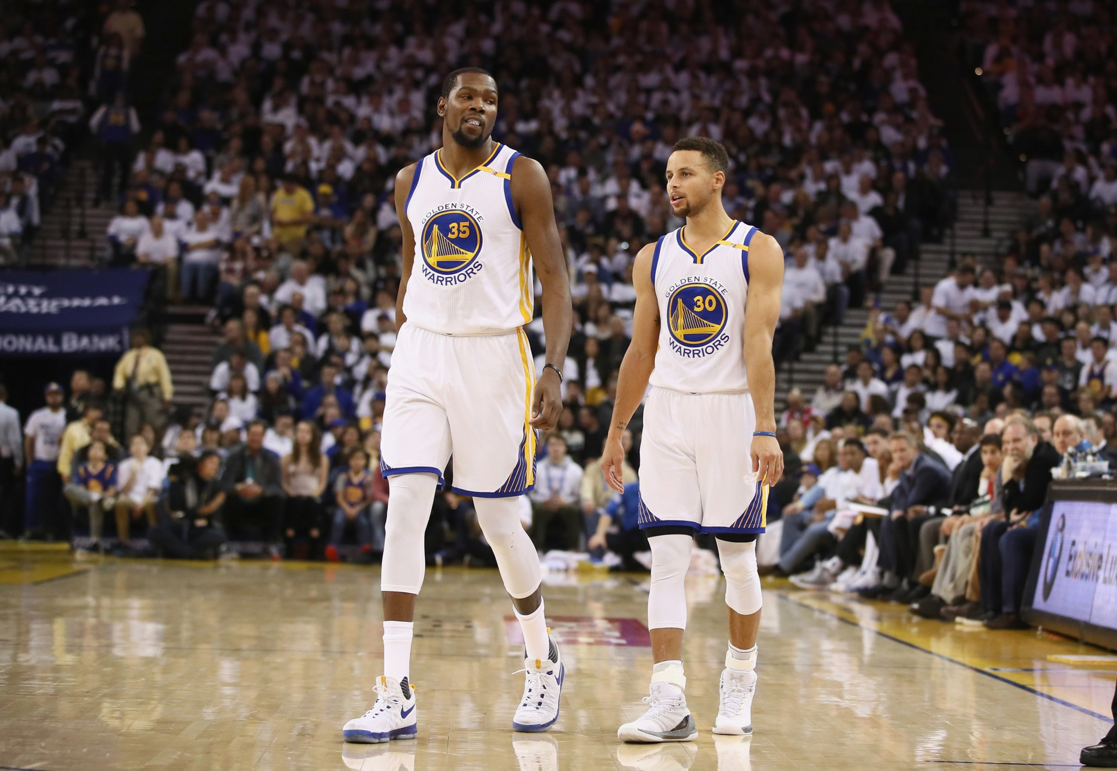 Two Golden State Warriors players in white uniforms, numbers 35 and 30, walk side by side on the basketball court during a game.