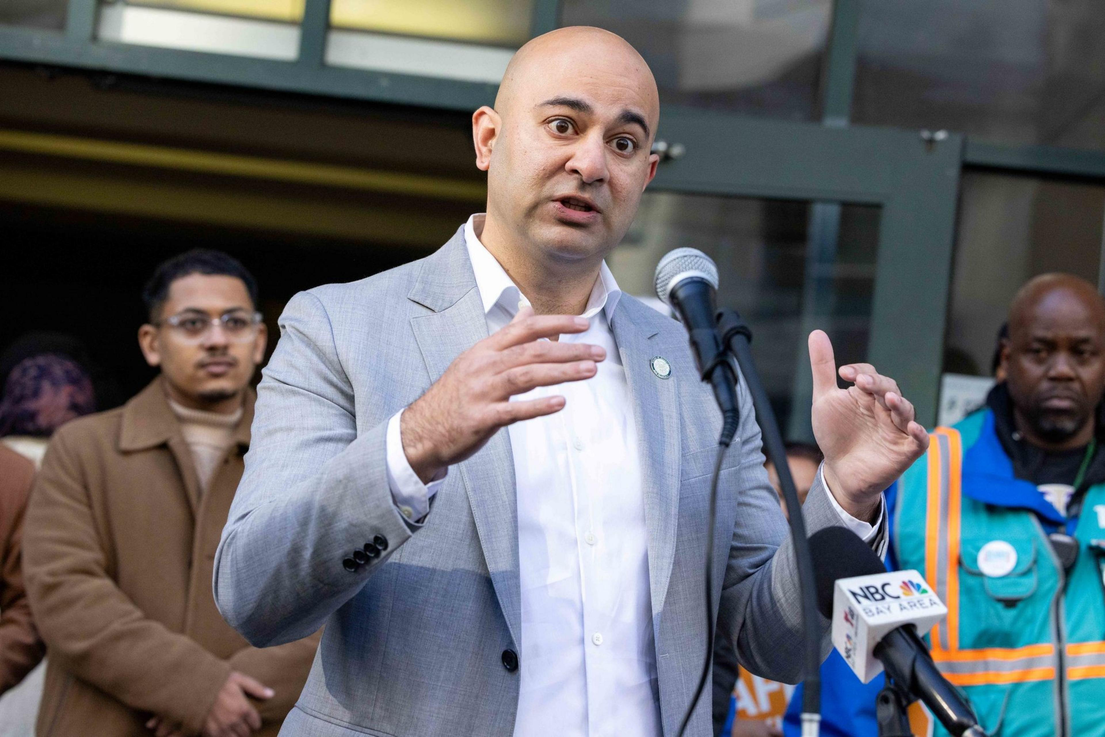 A man in a gray suit speaks passionately at a microphone with an &quot;NBC Bay Area&quot; sign. Two other men in the background observe him attentively.
