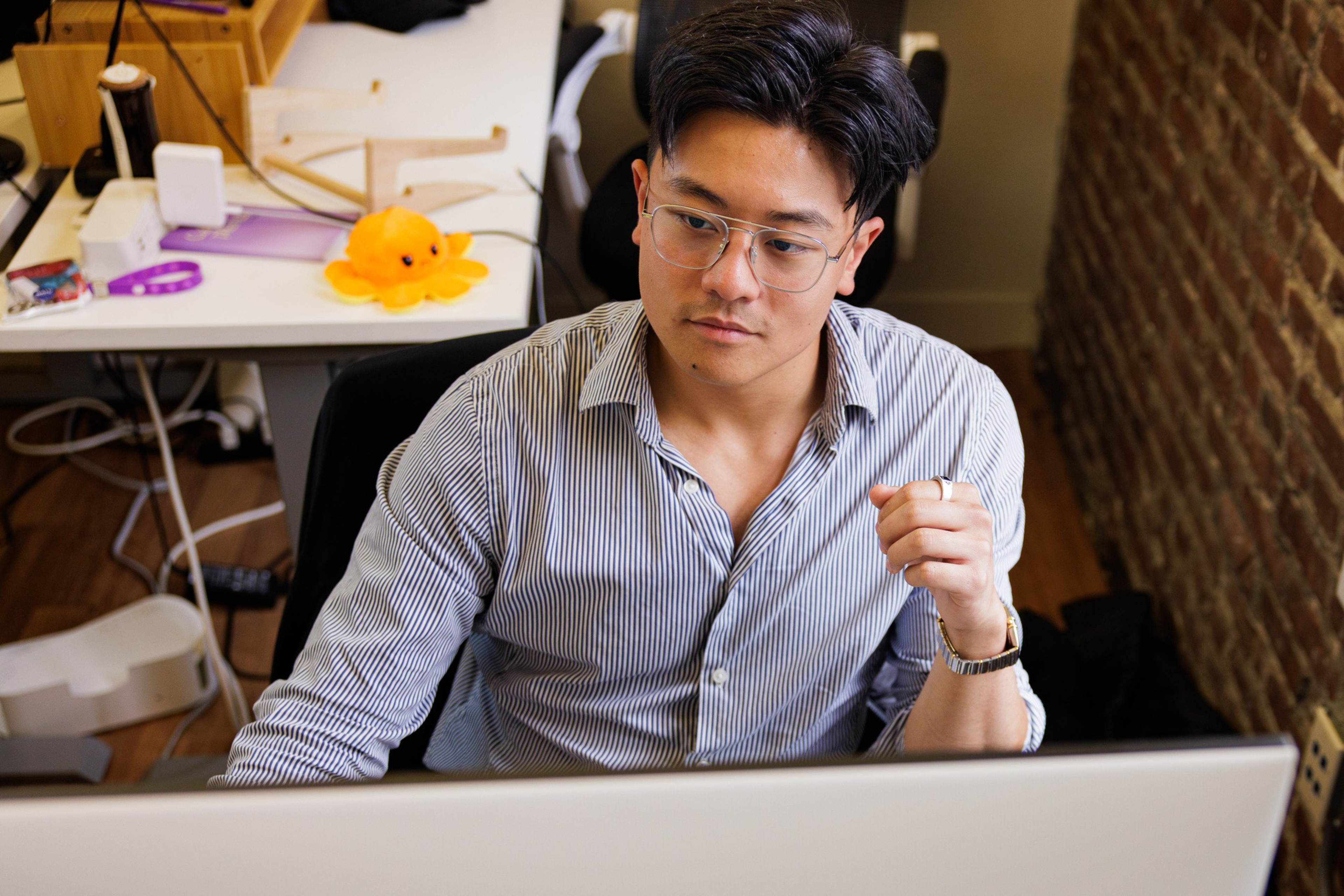 A man with glasses and a striped shirt is focused on a computer screen, sitting in a workspace with a brick wall and various items behind him.