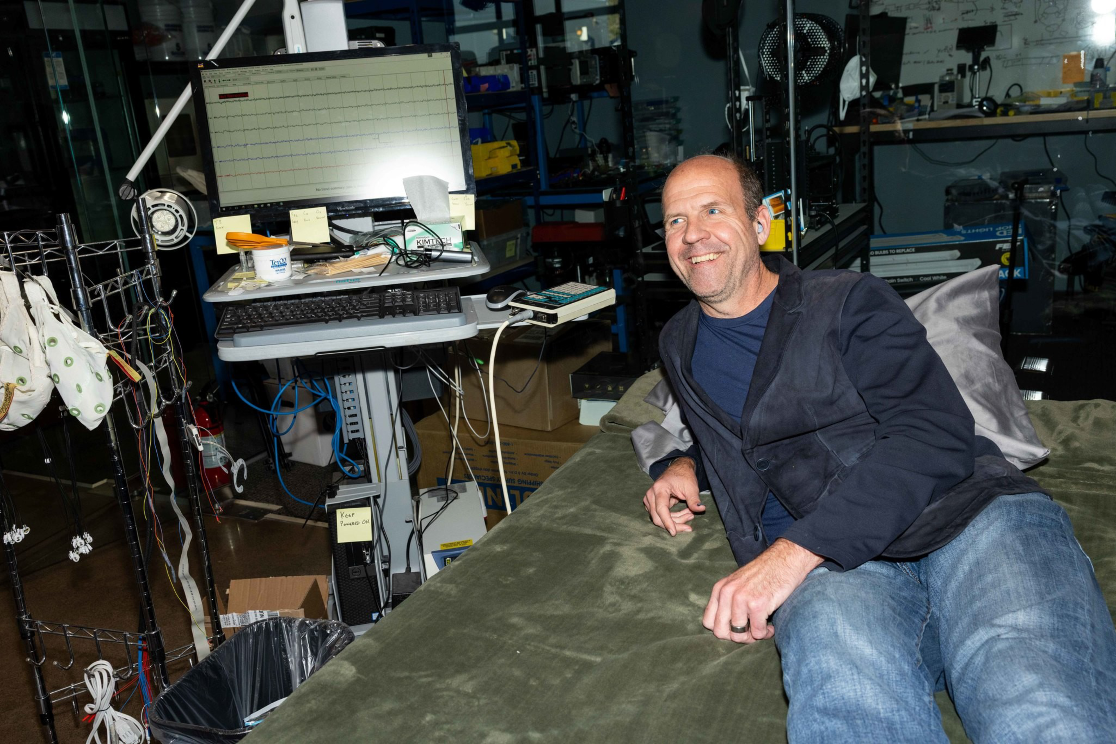 A man in casual clothes reclines on a green bed beside medical monitoring equipment displaying a heart rate graph.